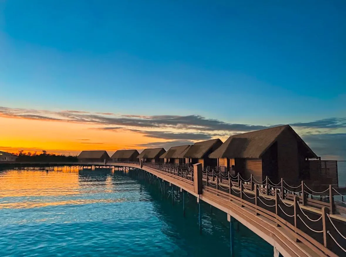A row of overwater bungalows on stilts extending over calm blue water at sunset with a clear sky.
