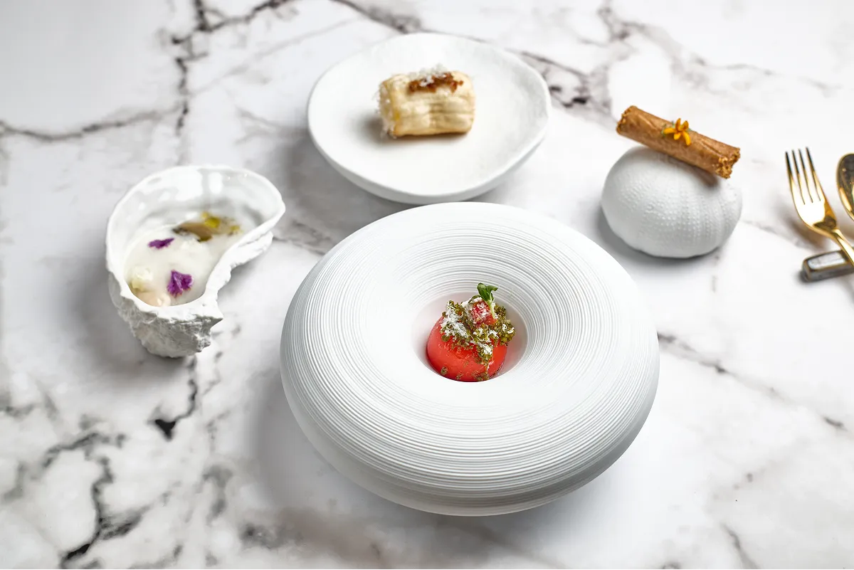 Artfully plated gourmet dish featuring a central white circular bowl with a red spherical element topped with herbs, accompanied by two white textured dishes with delicate food garnishes on a marble surface.