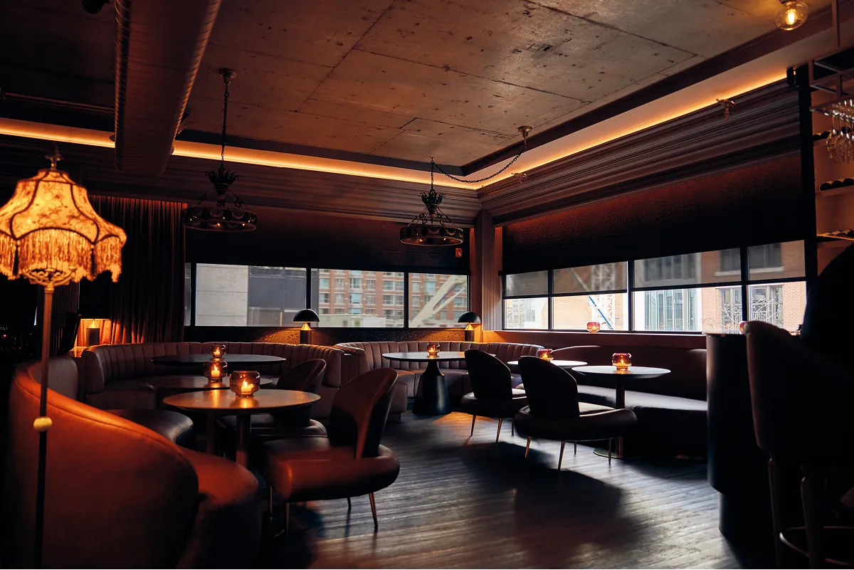Dimly lit modern lounge area with leather chairs, round tables, candles, and large windows overlooking city buildings.