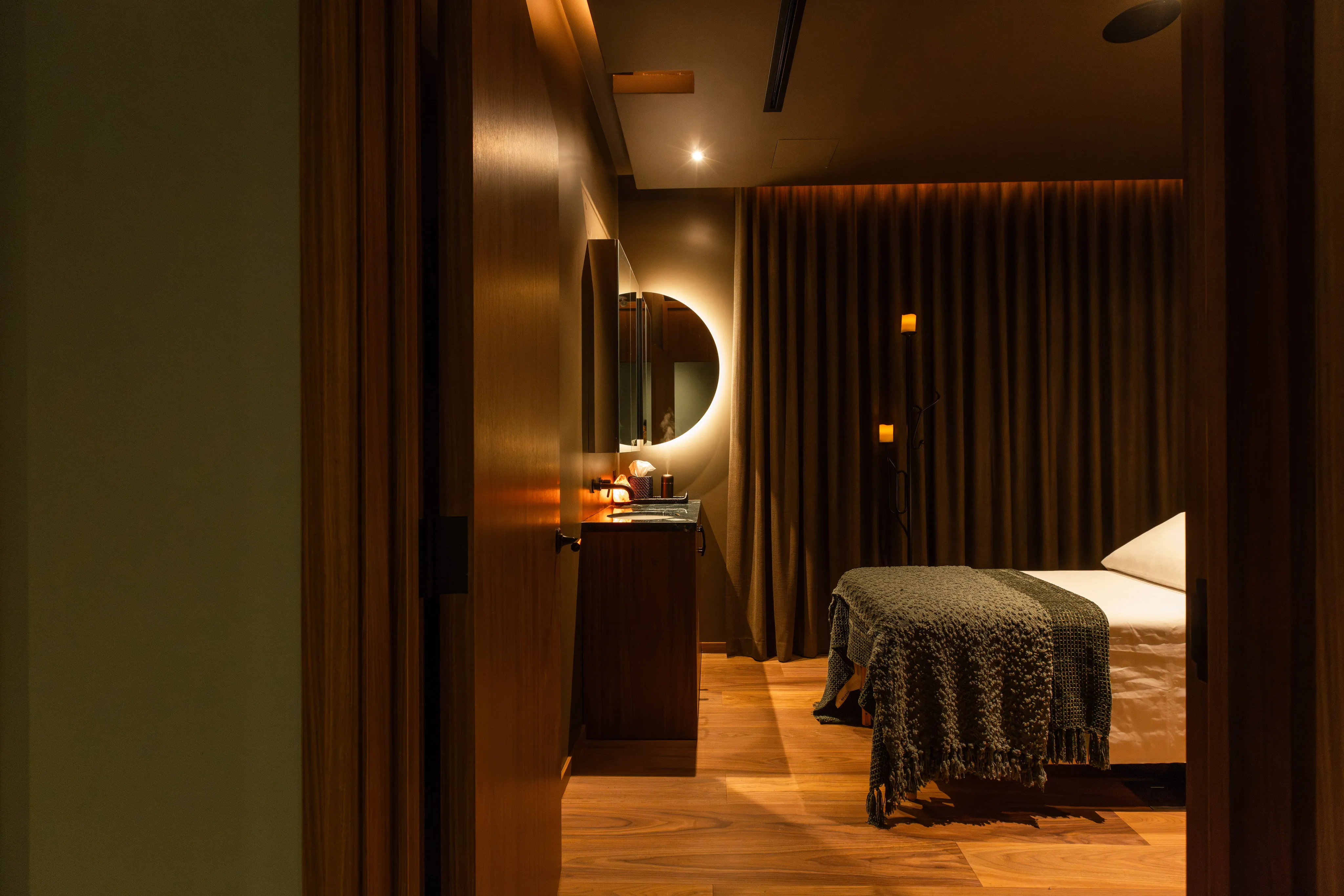Dimly lit massage room with wooden floor, a bed covered with a textured blanket, and a circular backlit mirror on the wall.