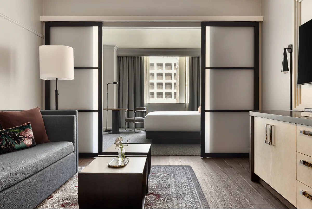 Modern hotel suite featuring a gray sofa, two nested coffee tables, sliding frosted glass doors, and a bedroom with a large window in the background.