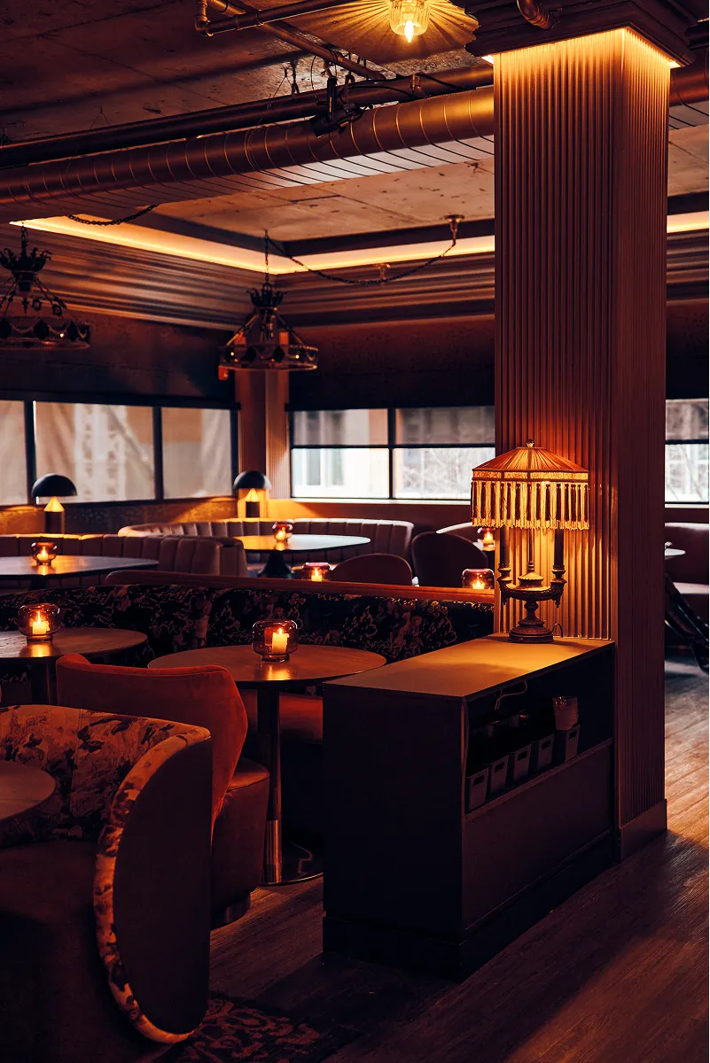 Cozy dimly lit lounge with velvet chairs, round tables, candle lights, and vintage lamp on a sideboard.
