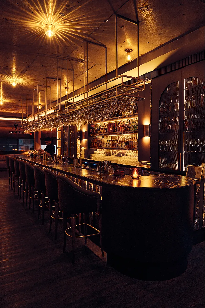 Dimly lit upscale bar with a long marble counter, hanging wine glasses, and a bartender preparing drinks.