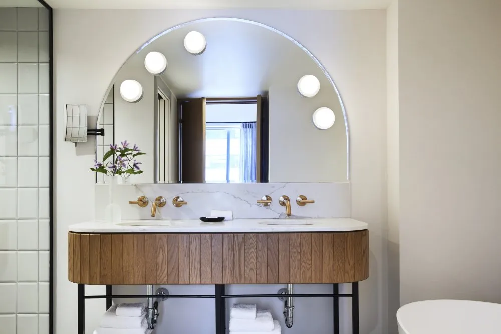 Modern bathroom vanity with double sinks, brass faucets, round illuminated mirror, wooden cabinet, and white tiled wall.