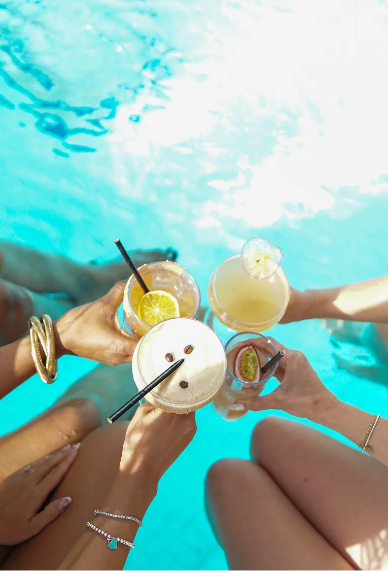 Four people holding colorful cocktails over a bright blue pool.