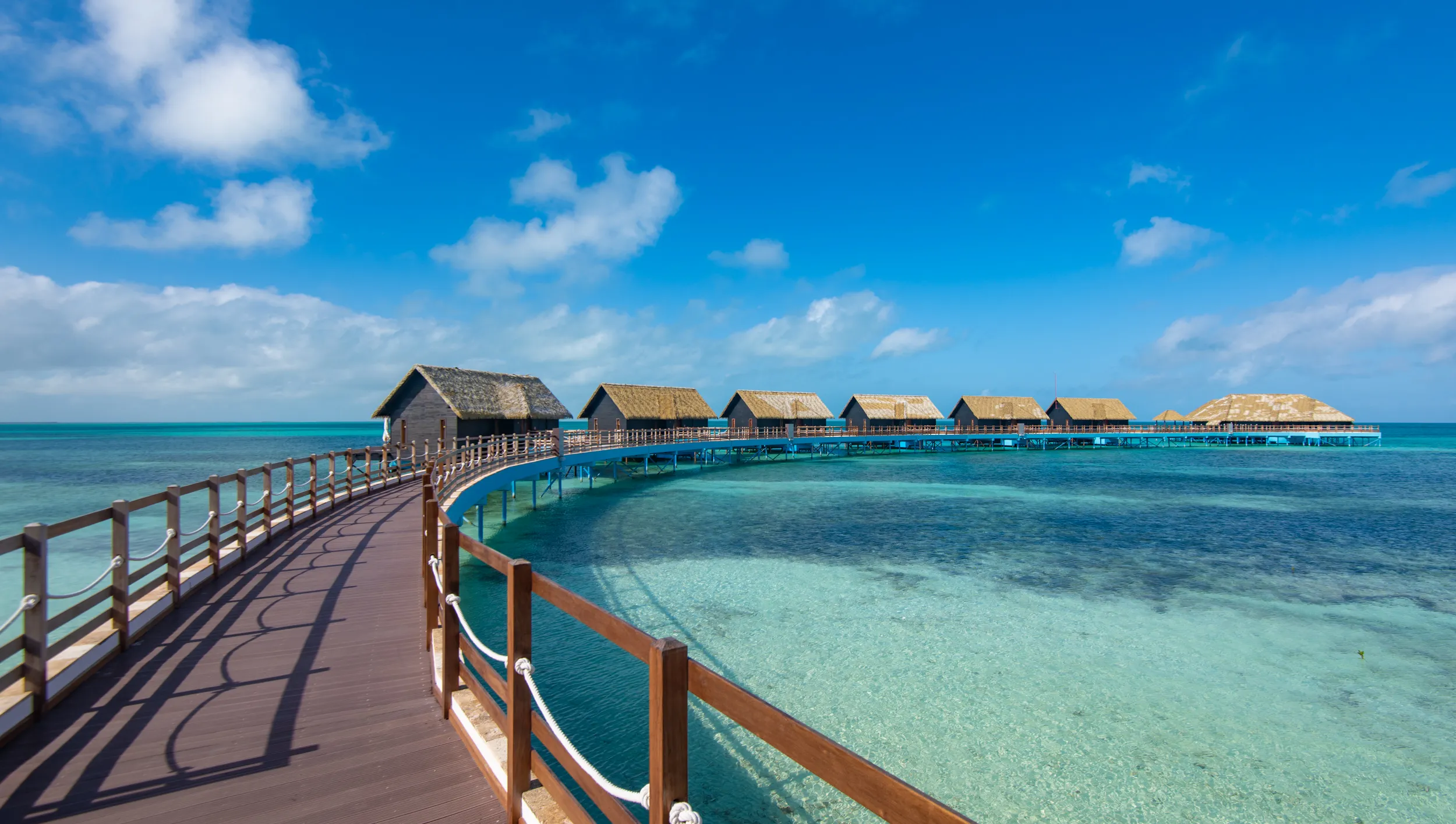 Wooden walkway curving over clear turquoise water leading to a series of overwater bungalows under a blue sky with scattered clouds.