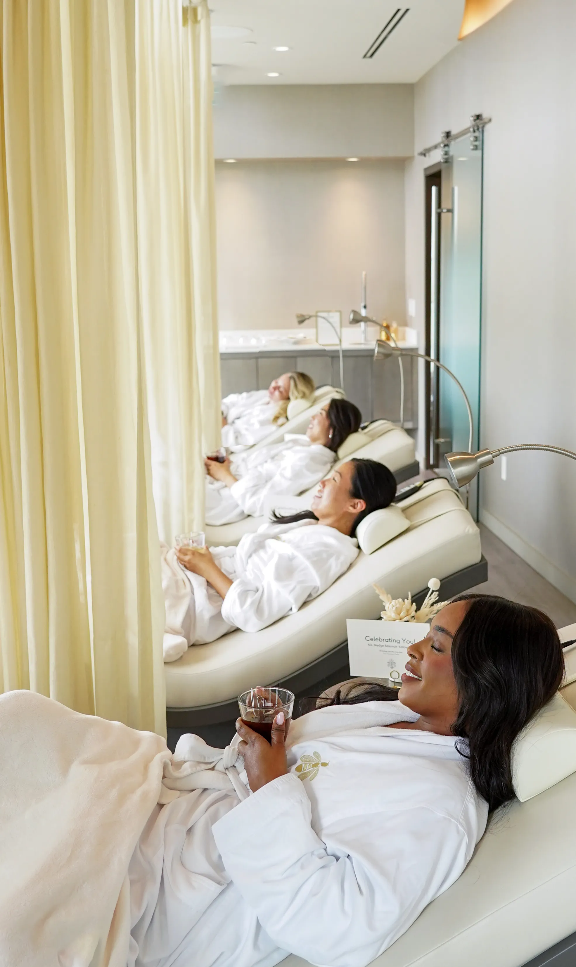 Four women in white robes relaxing on lounge chairs with drinks in a spa room separated by a cream curtain.