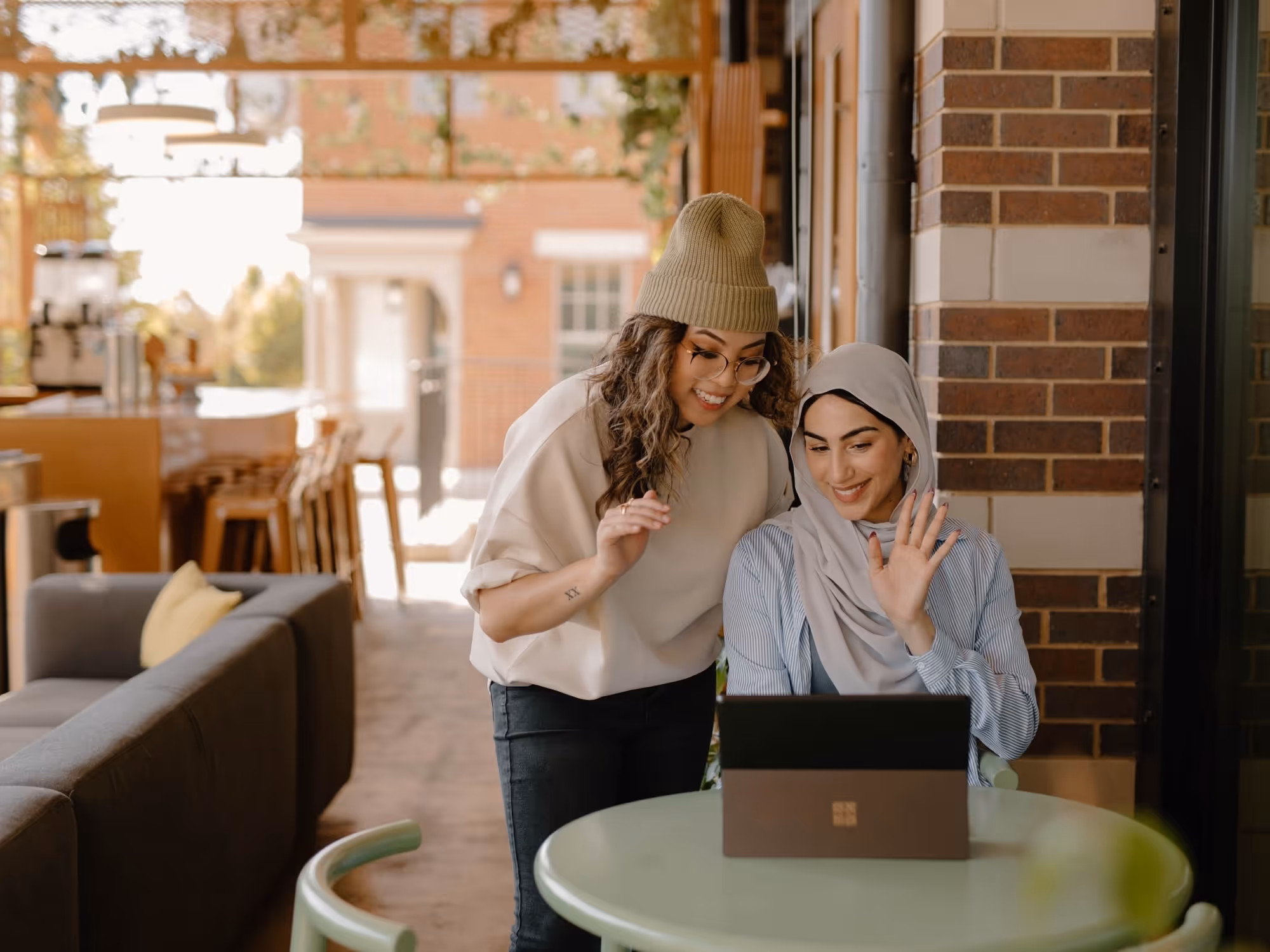 Two women smiling and waving at a laptop screen while sitting at a round table in a cozy indoor space.