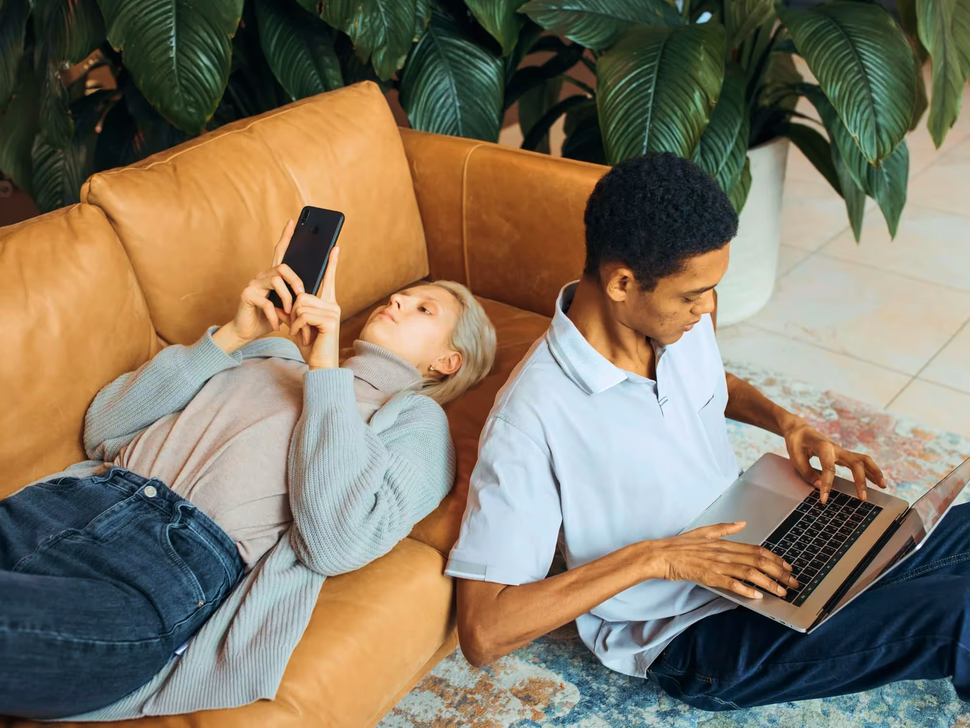 A woman lying on a brown leather couch looking at her smartphone and a man sitting nearby on the floor using a laptop, with large green plants in the background.