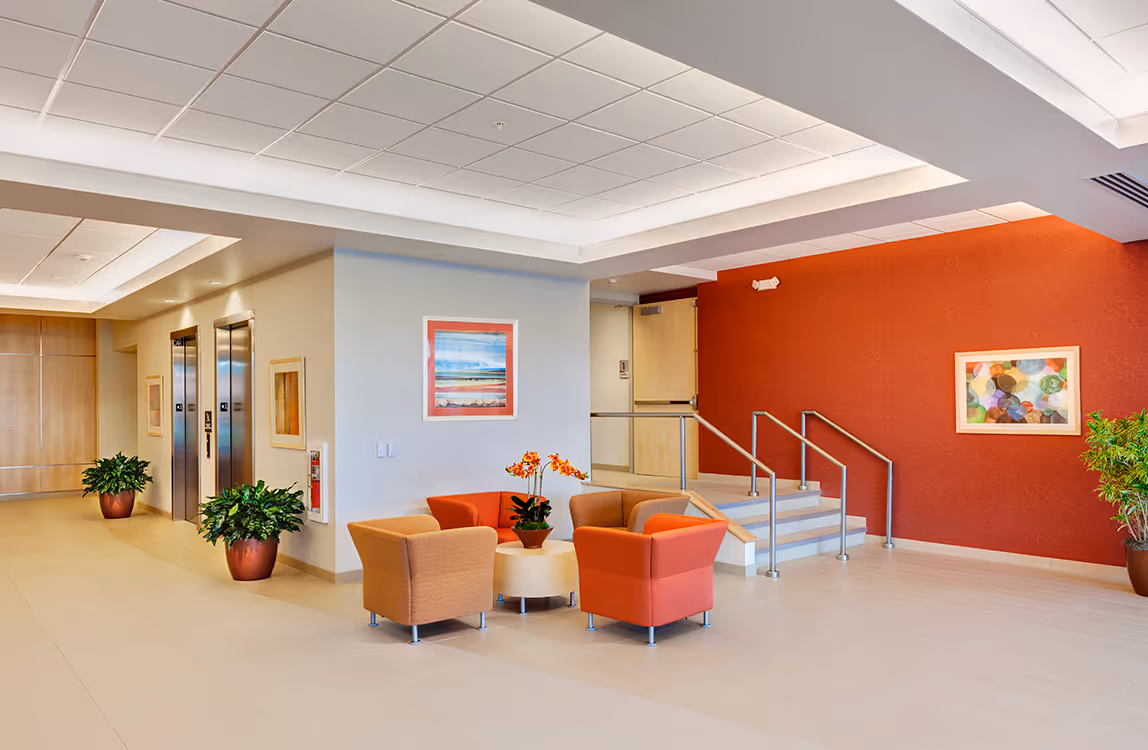 Modern lobby area with a seating arrangement of four chairs around a small round table with an orange orchid, two elevators on the left, red accent wall with framed artwork, and potted plants.