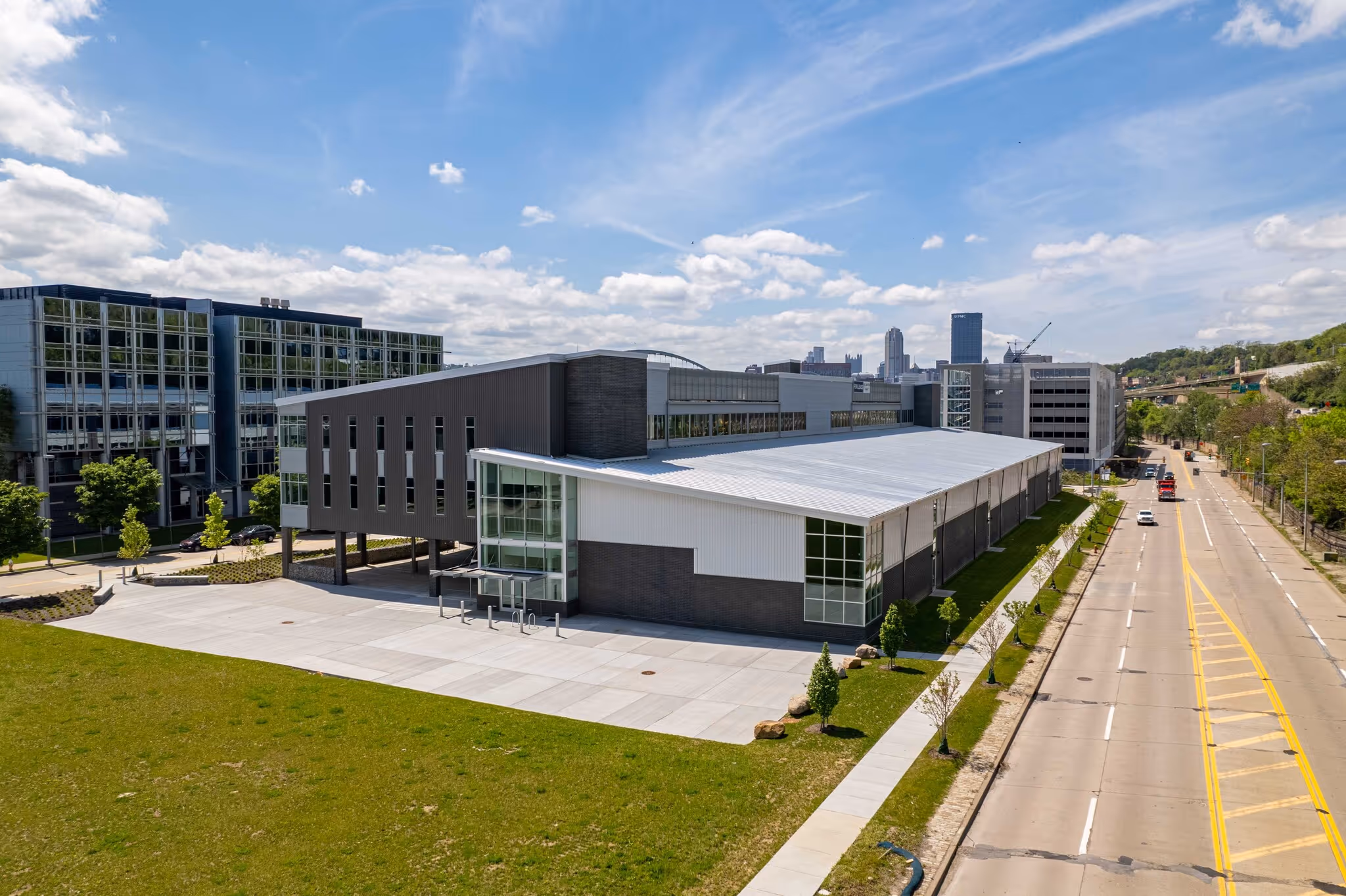 Modern commercial building with metal and glass facade beside a street lined with young trees under a blue sky with scattered clouds.