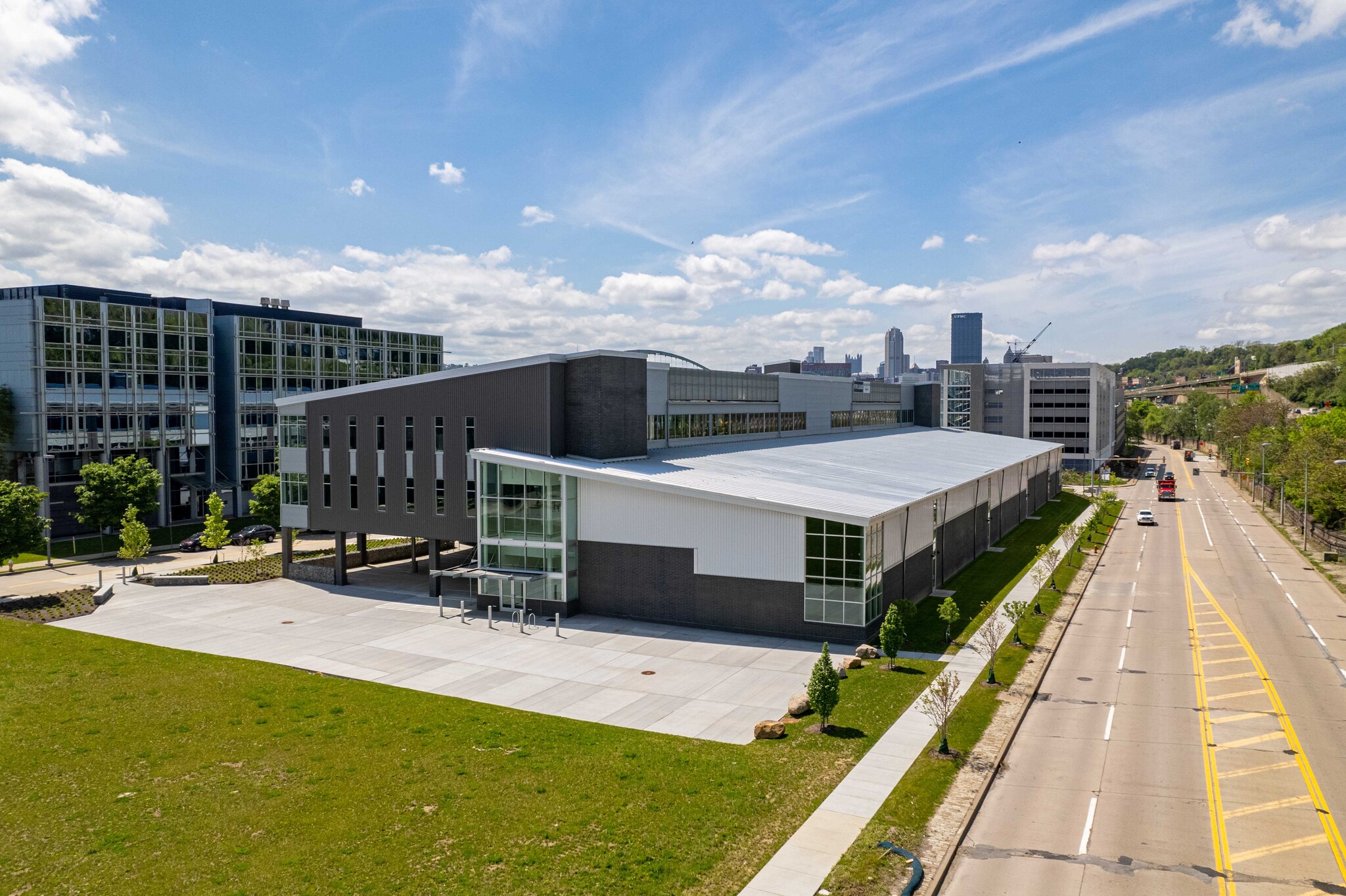 Modern commercial building with metal and glass facade beside a street lined with young trees under a blue sky with scattered clouds.