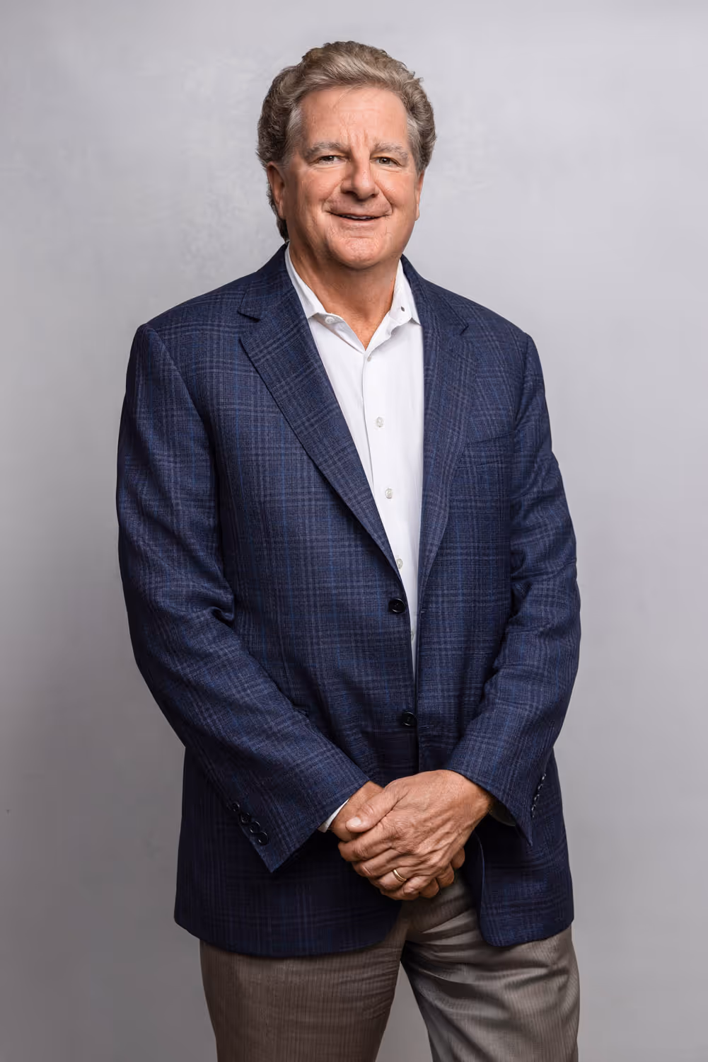 Smiling middle-aged man in a blue plaid blazer and white shirt, standing with hands clasped.