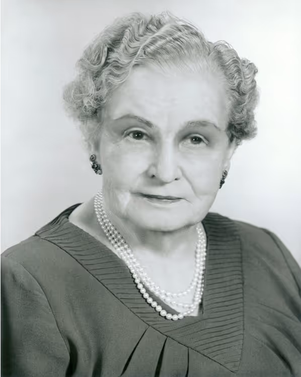 Black and white portrait of an elderly woman with curled hair wearing a dark dress and a double-strand pearl necklace.
