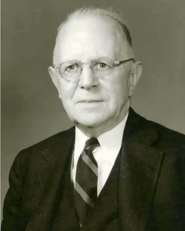 Black and white portrait of an older man wearing glasses, a dark suit, white shirt, and striped tie.