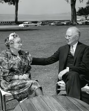 Black and white photo of an elderly man and woman seated outdoors on lawn chairs, the man gently touching the woman's shoulder.