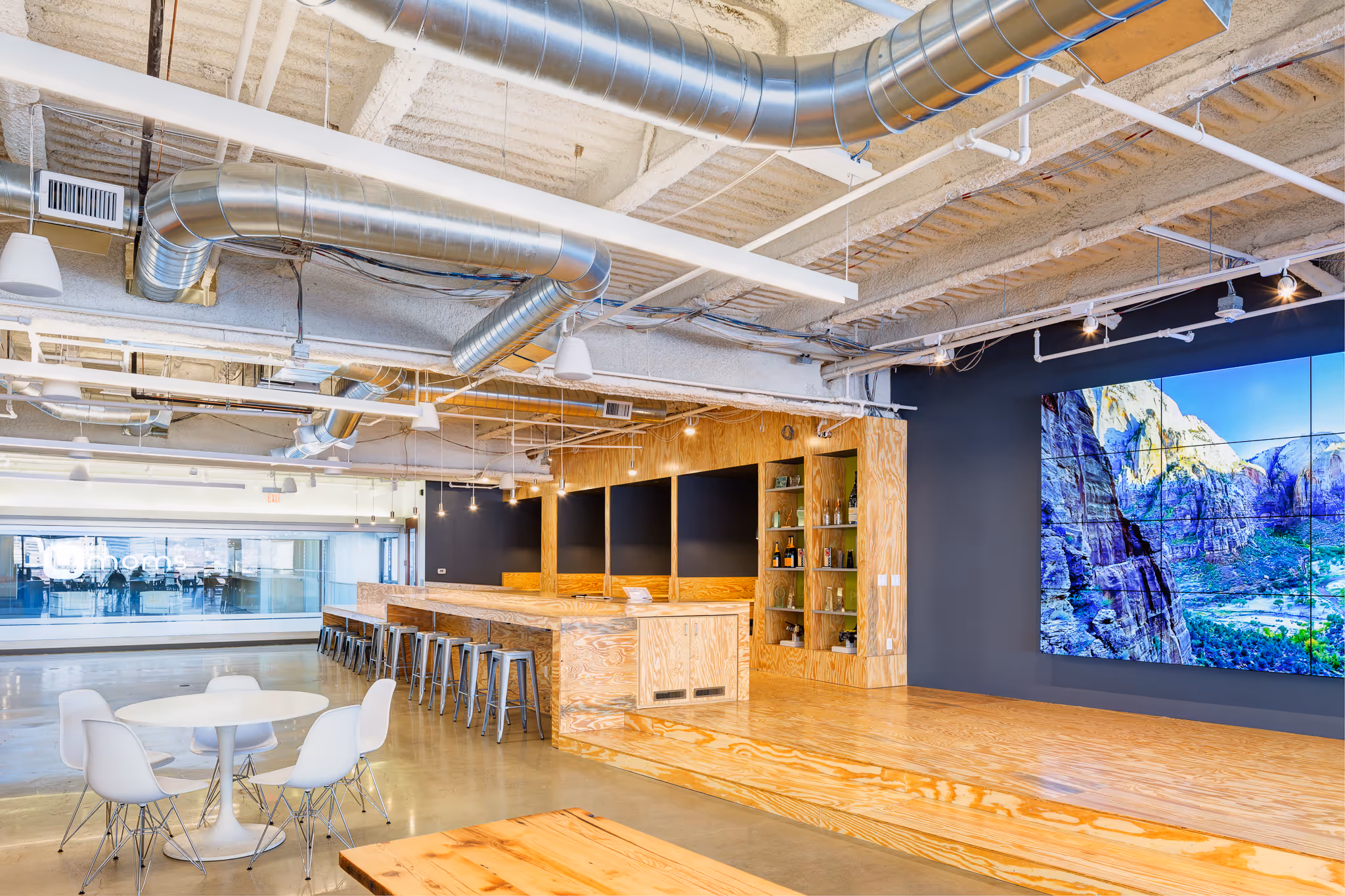 Modern office lounge with wooden seating area, metal stools, white round table with chairs, and a large screen displaying a mountain landscape.