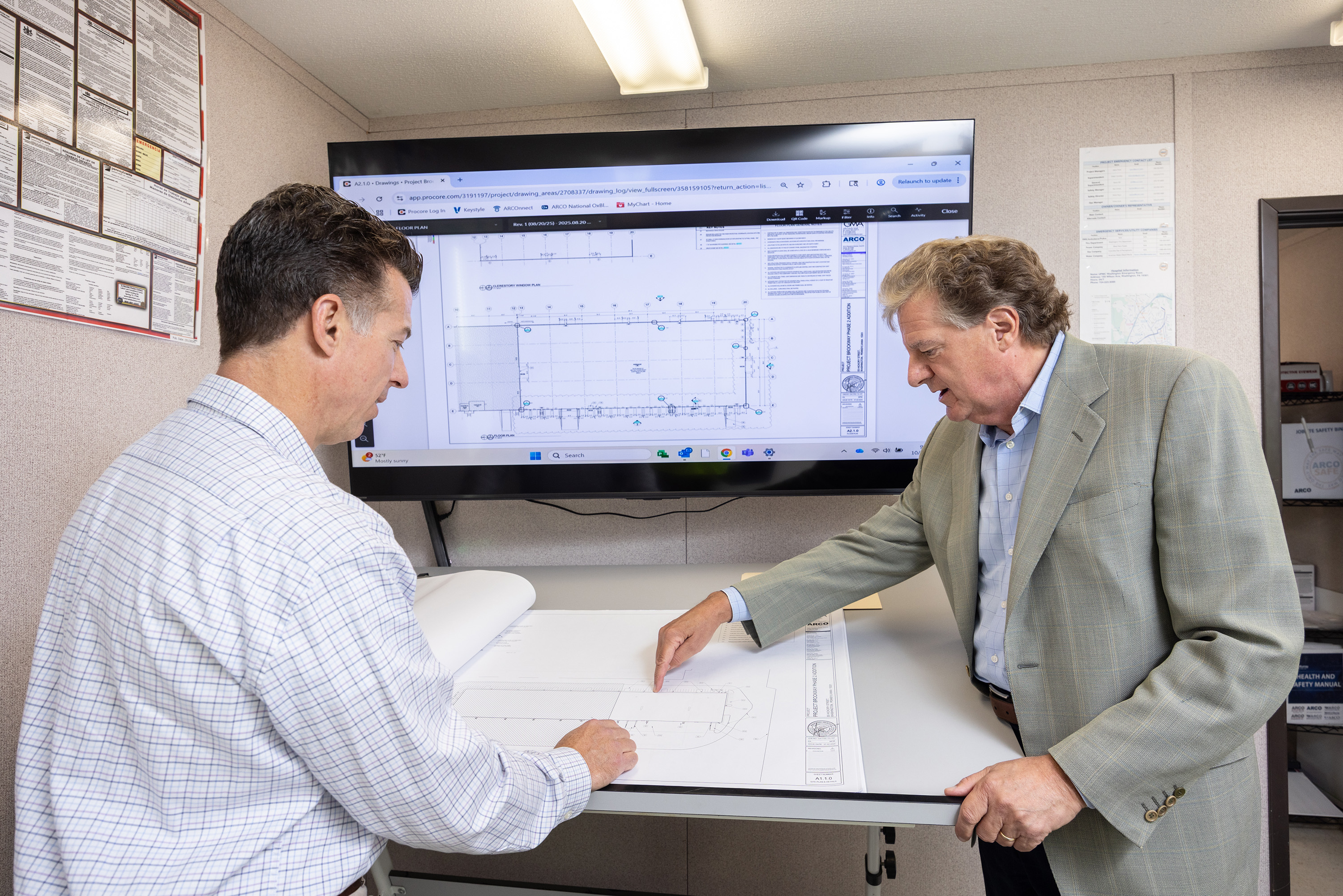 Two men reviewing architectural drawings on a table with the same plan displayed on a large screen behind them in an office.