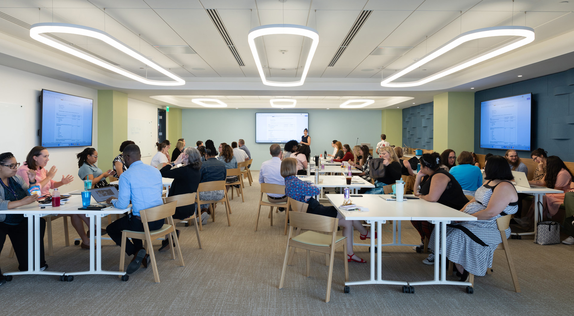 A diverse group of adults attending a classroom-style training session with a presenter at the front and multiple large monitors displaying information.