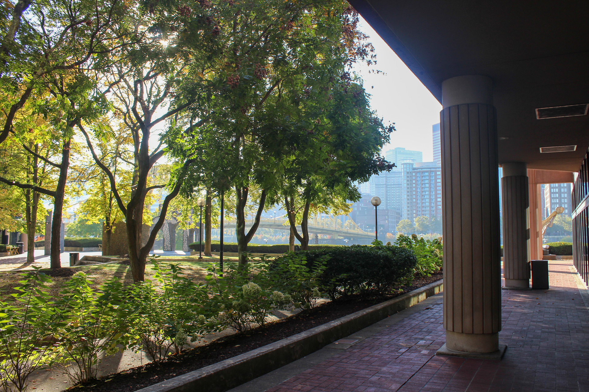 Sunlit park area with green trees, bushes, and pathway next to tall building columns, city skyline and a bridge in the background.