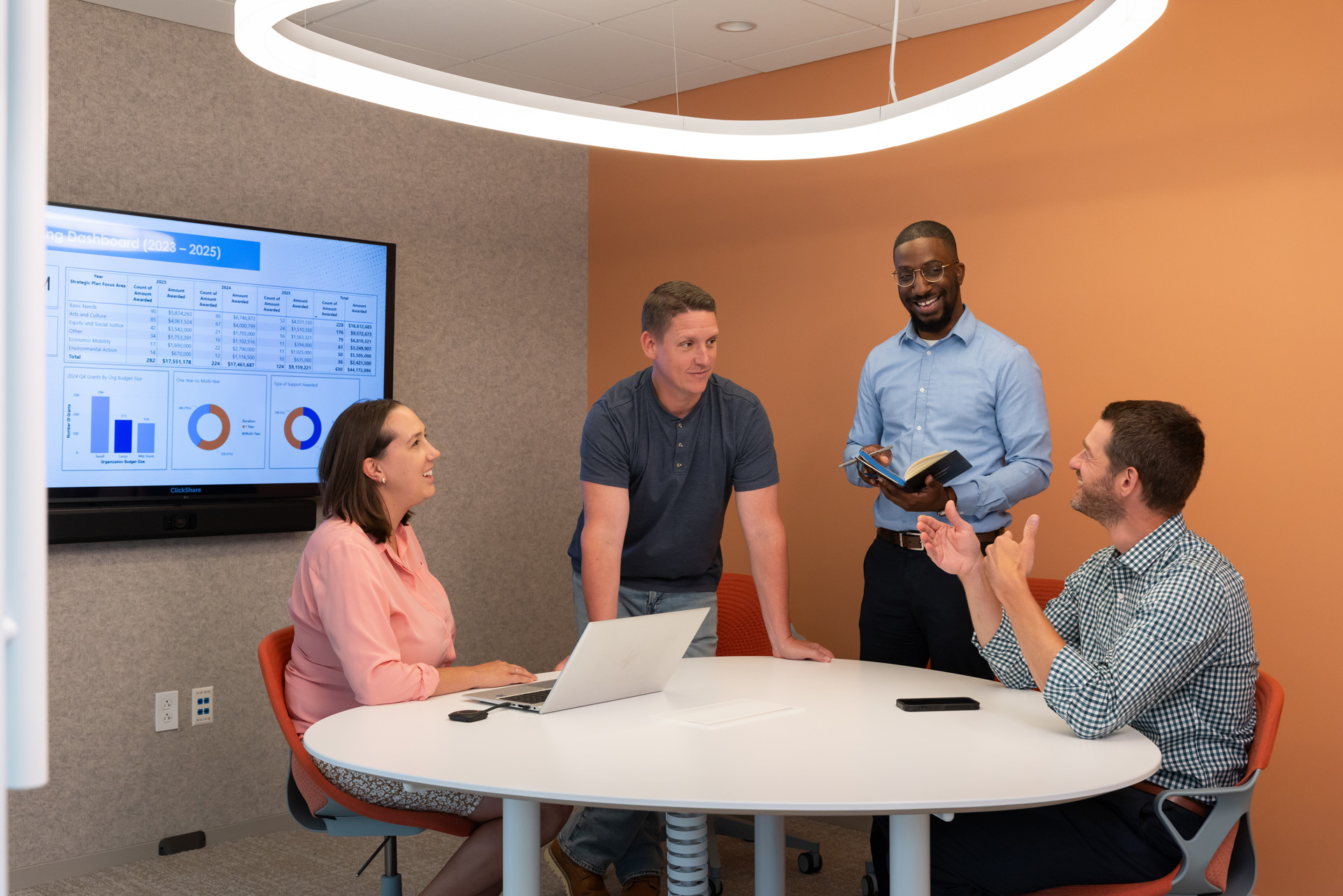 Four colleagues in a meeting room discuss data shown on a screen; one person stands with a notebook while others sit around a white round table.