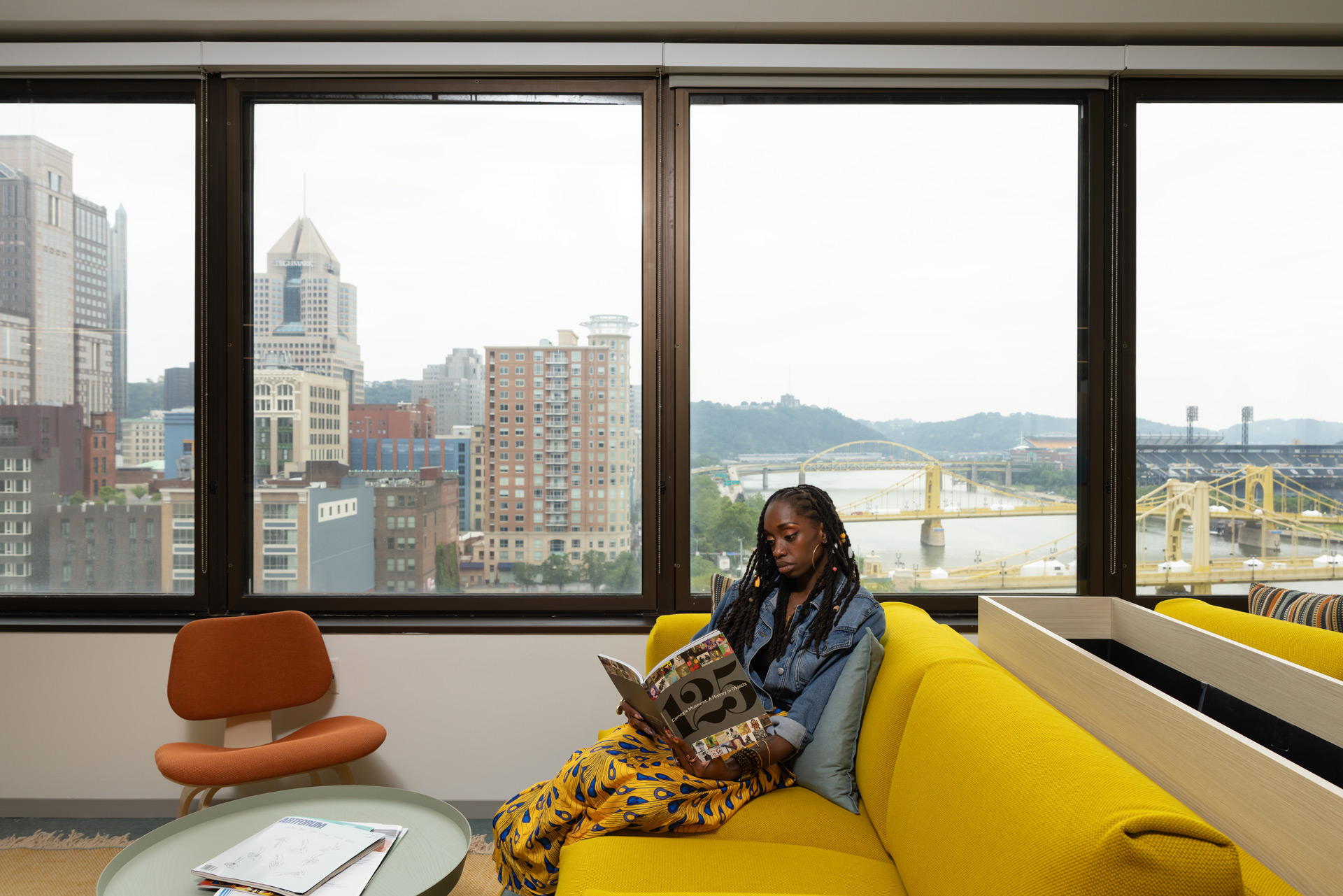 Woman with braided hair reading a magazine while sitting on a bright yellow couch in a room with large windows showing a city skyline and yellow bridge.