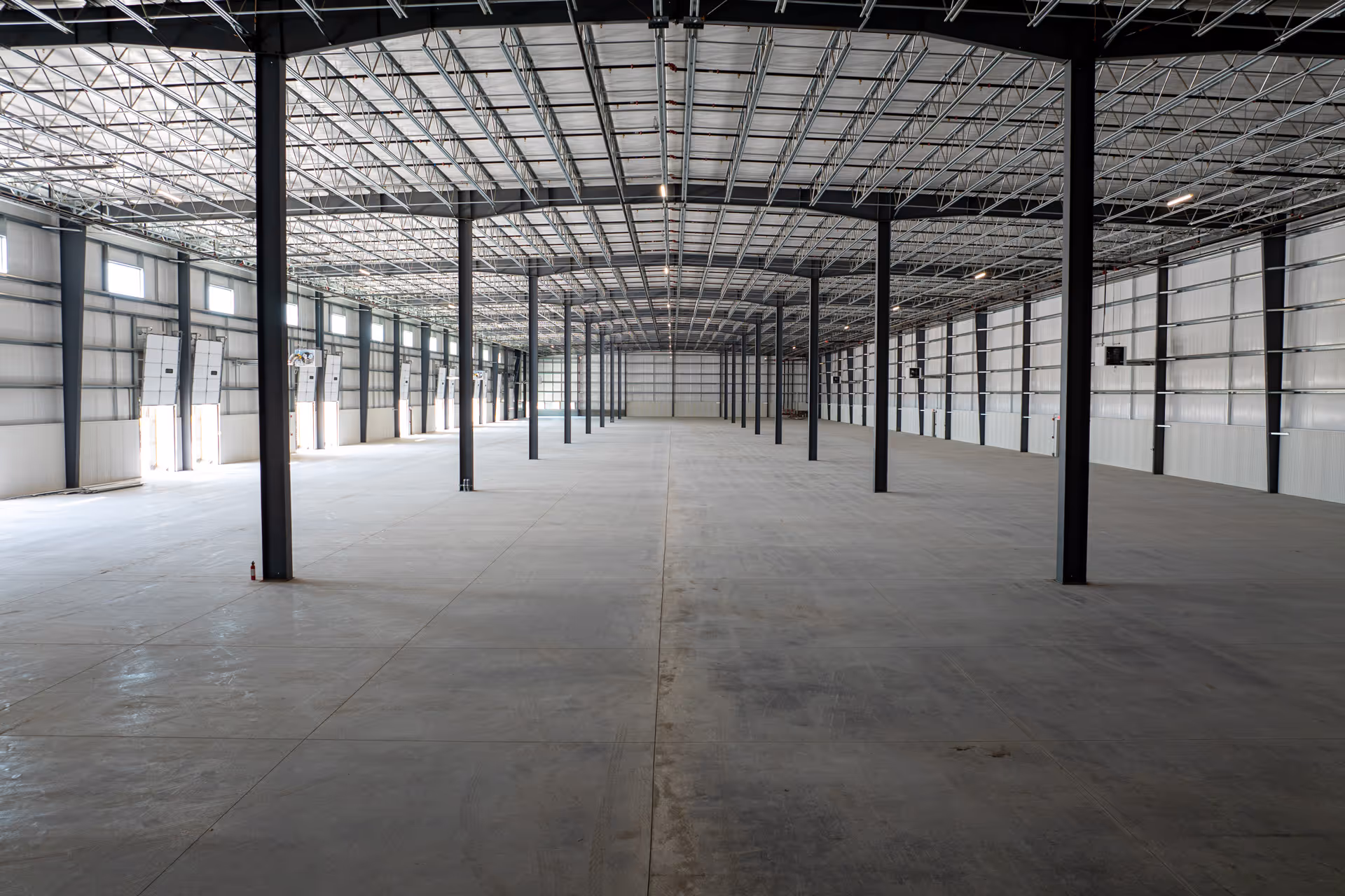 Spacious empty industrial warehouse with metal support beams and concrete floors under a metal truss ceiling.
