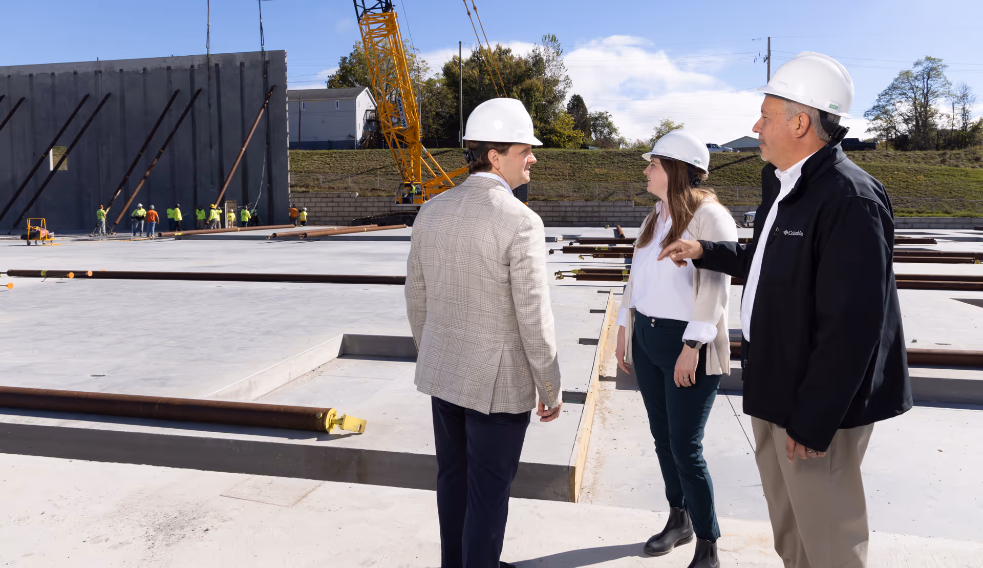 Three construction professionals wearing hard hats discussing at a building site with workers and crane in the background.