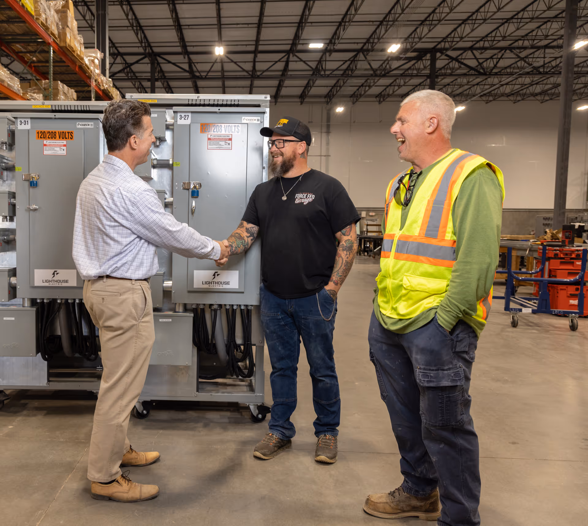 Three men in a warehouse, one in business attire shaking hands with a tattooed man in casual wear, while a man in a high-visibility vest smiles nearby.