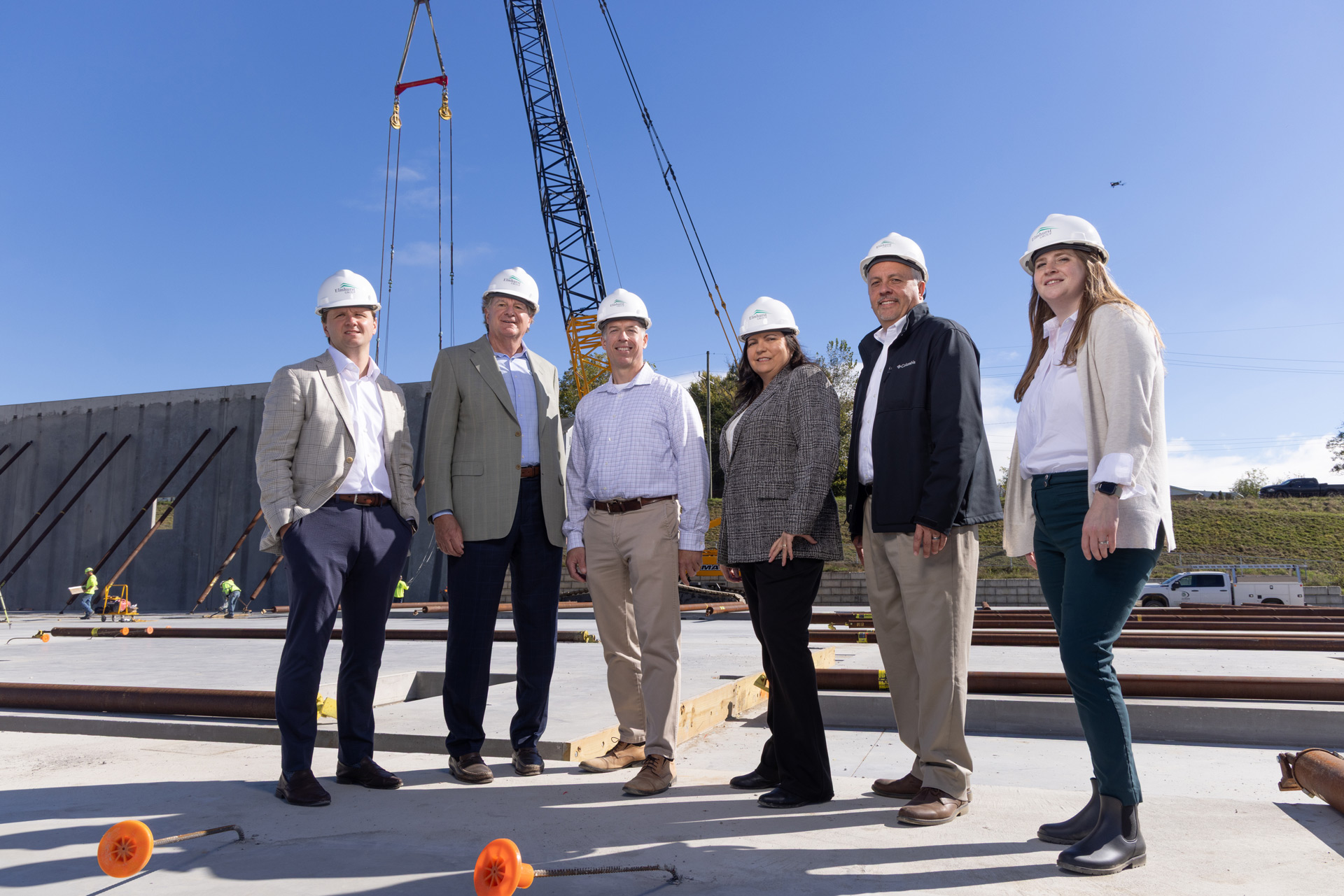 Six construction professionals wearing hard hats standing on a construction site with a crane and unfinished concrete walls in the background under a clear blue sky.