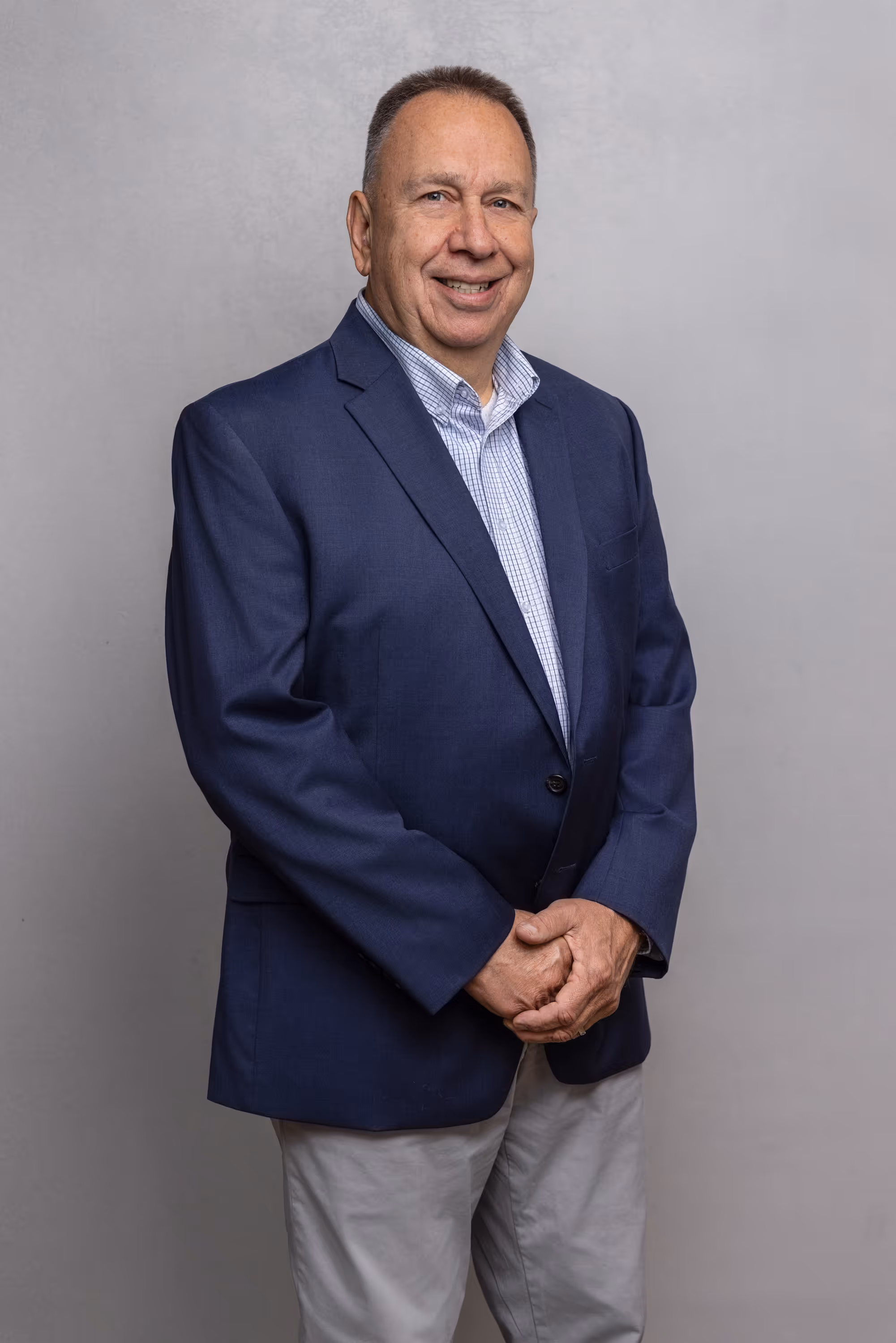 Smiling middle-aged man in a navy blazer, light checkered shirt, and light-colored pants standing against a gray background.