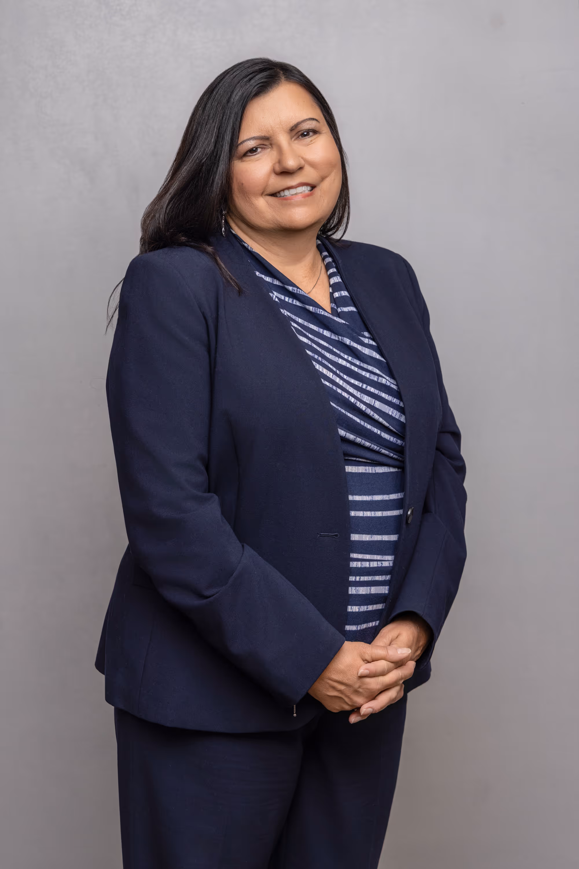 Smiling woman with long dark hair wearing a navy blazer and striped blouse, standing with hands clasped in front.