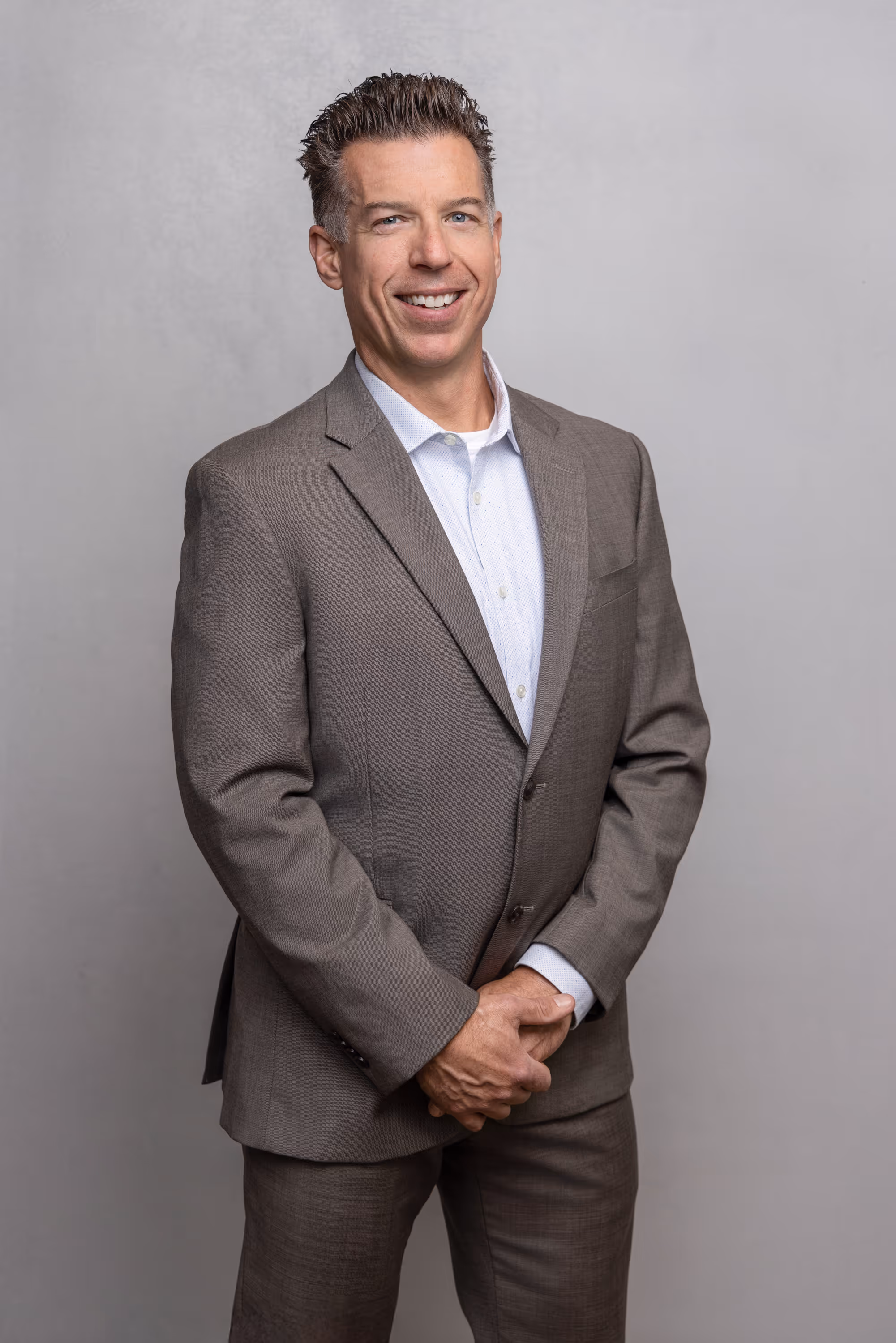 Smiling man in a brown suit and white shirt standing against a plain gray background.