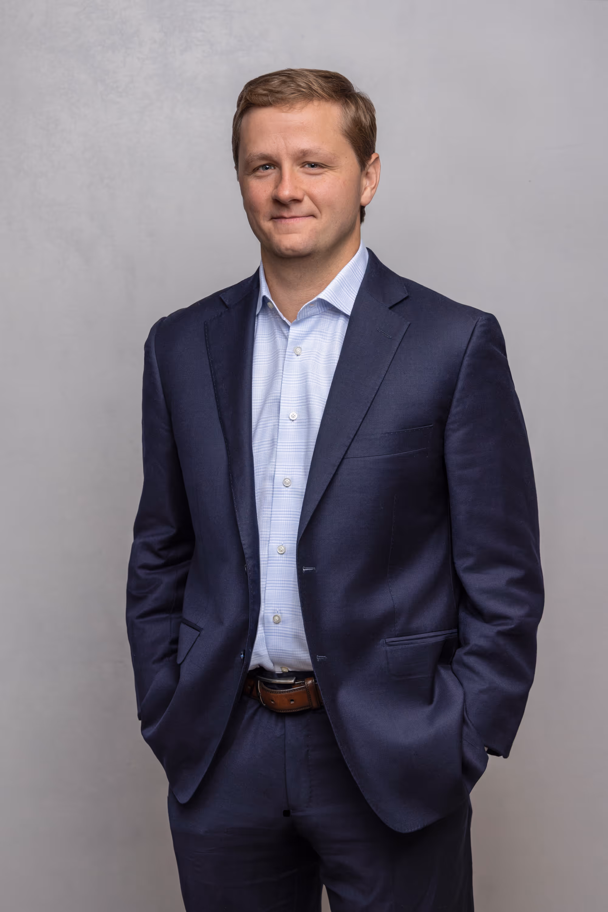 Man wearing a navy blue suit and light blue checkered shirt standing with hands in pockets against a light gray background.
