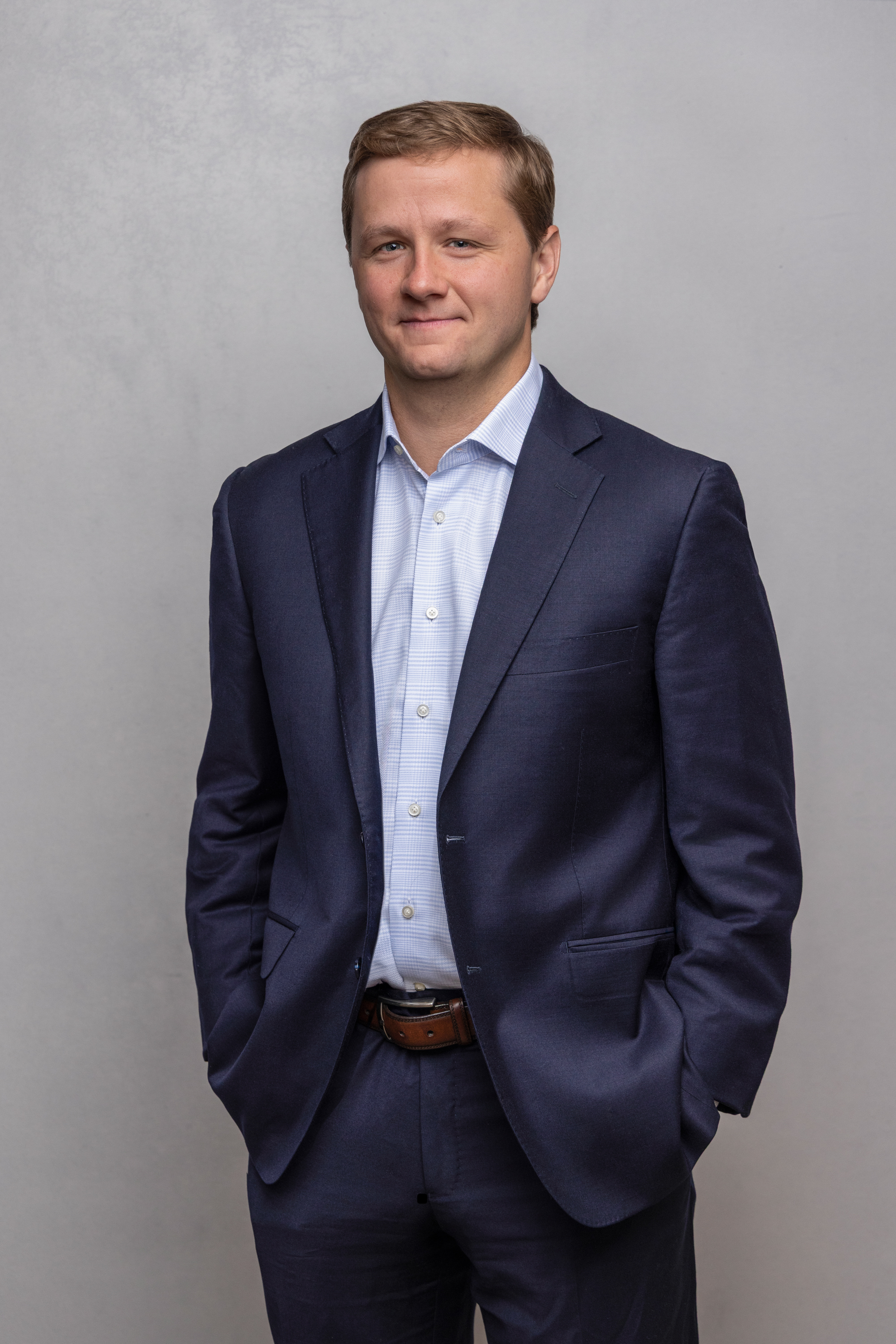 Man wearing a navy blue suit and light blue checkered shirt standing with hands in pockets against a light gray background.