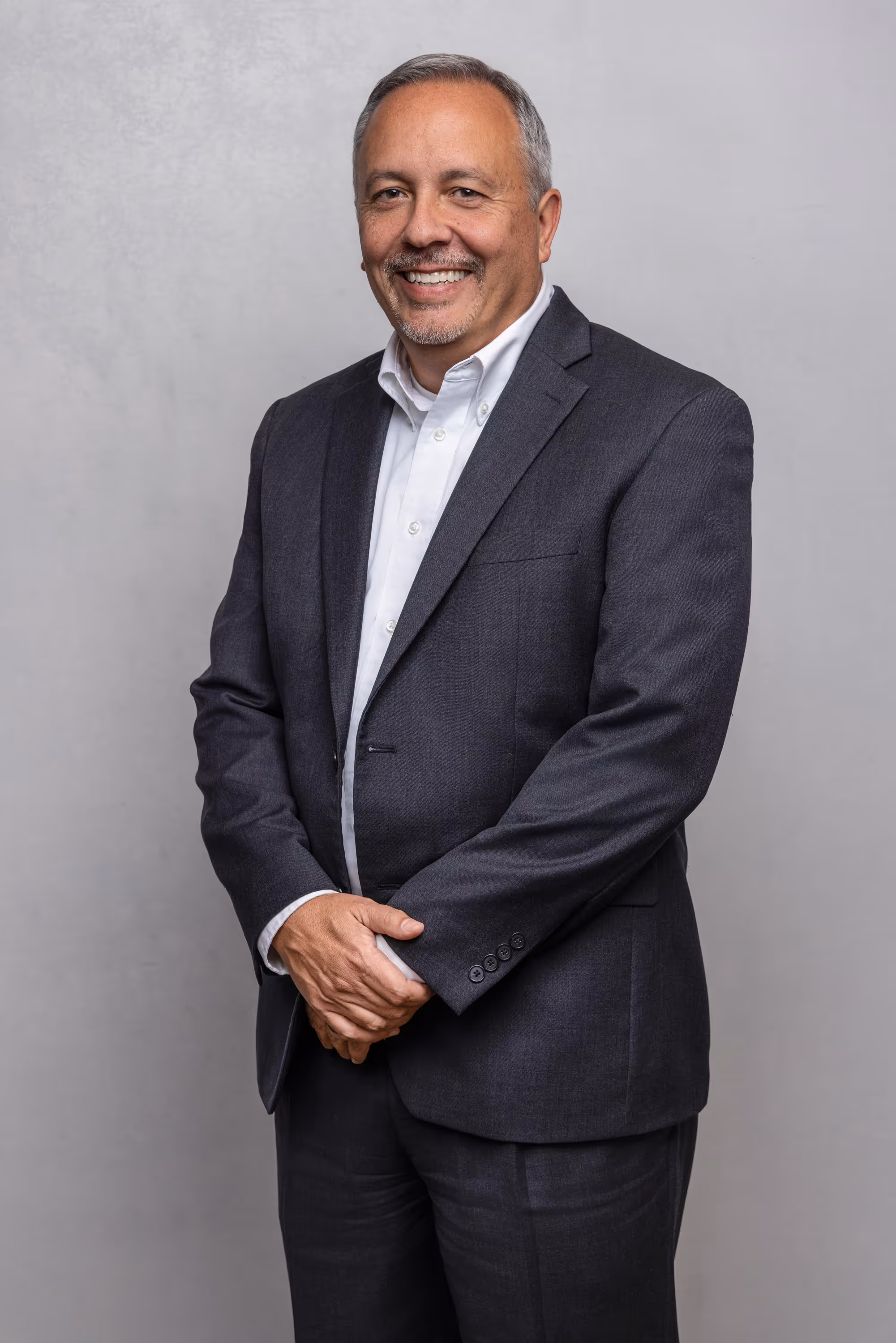 Smiling middle-aged man wearing a dark gray suit and white shirt standing against a light gray background.