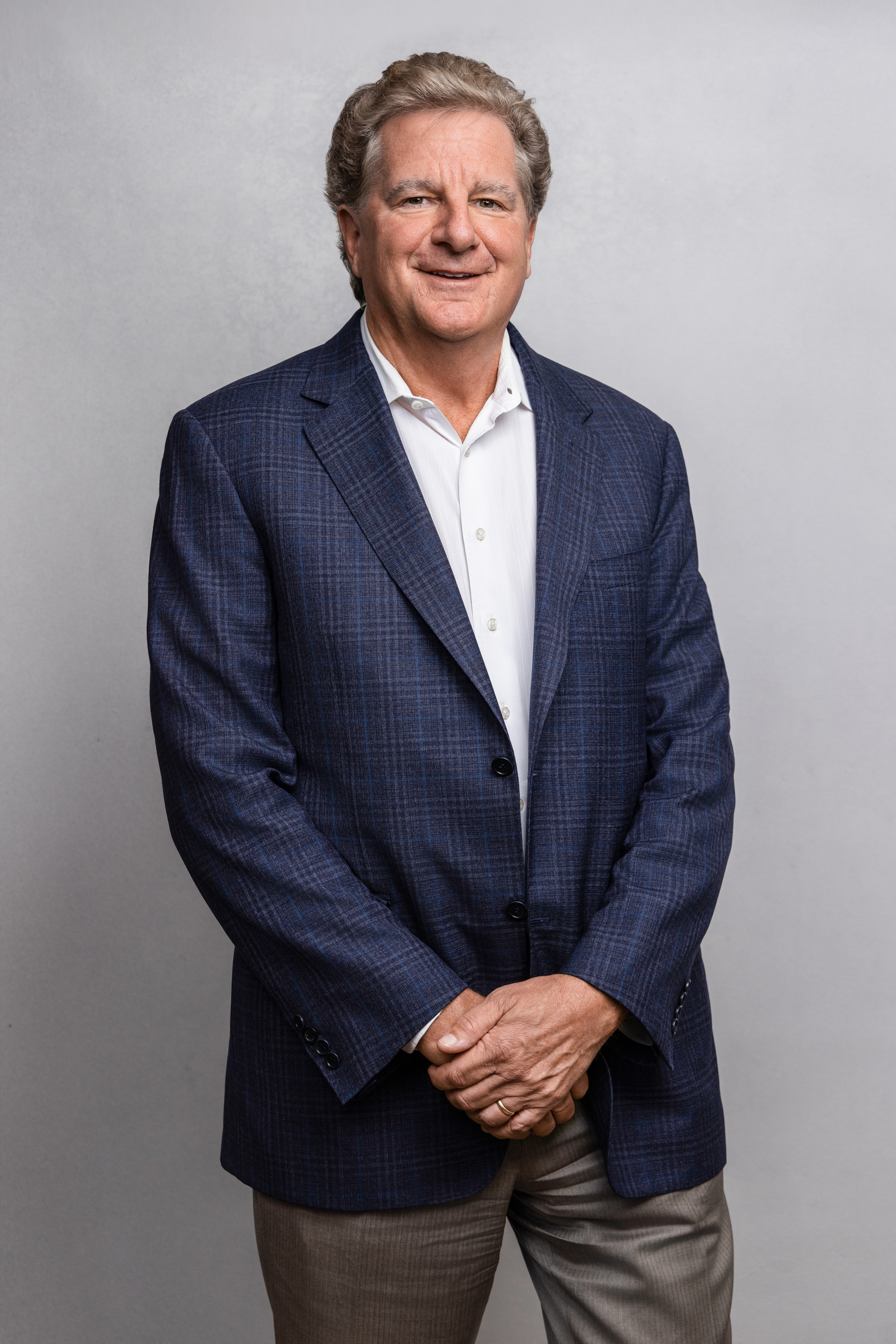 Middle-aged man with short curly hair wearing a blue plaid blazer, white dress shirt, and brown pants, standing against a light gray background.