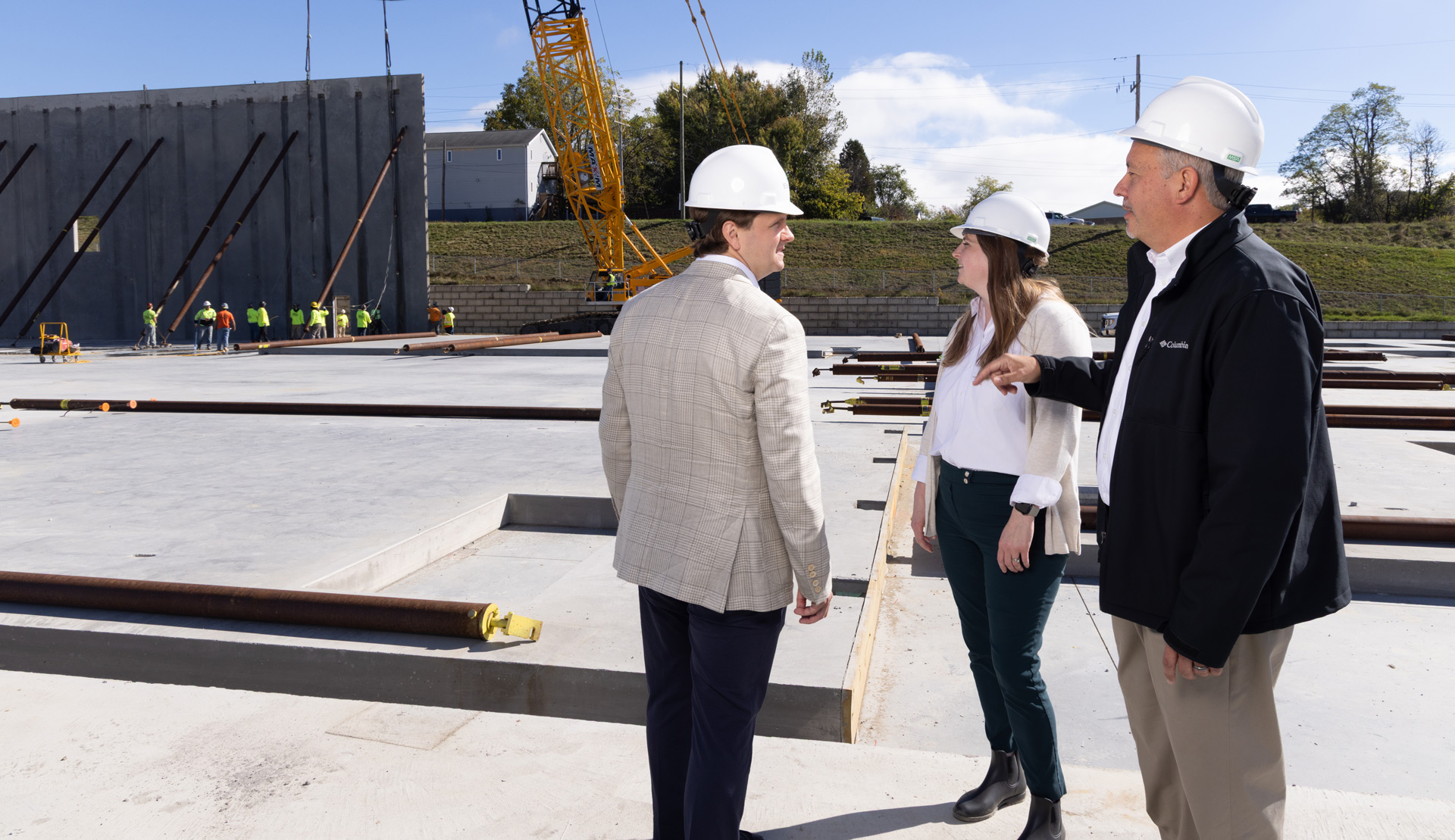 Three construction professionals wearing white hard hats are discussing at a construction site with a concrete foundation and workers in the background.