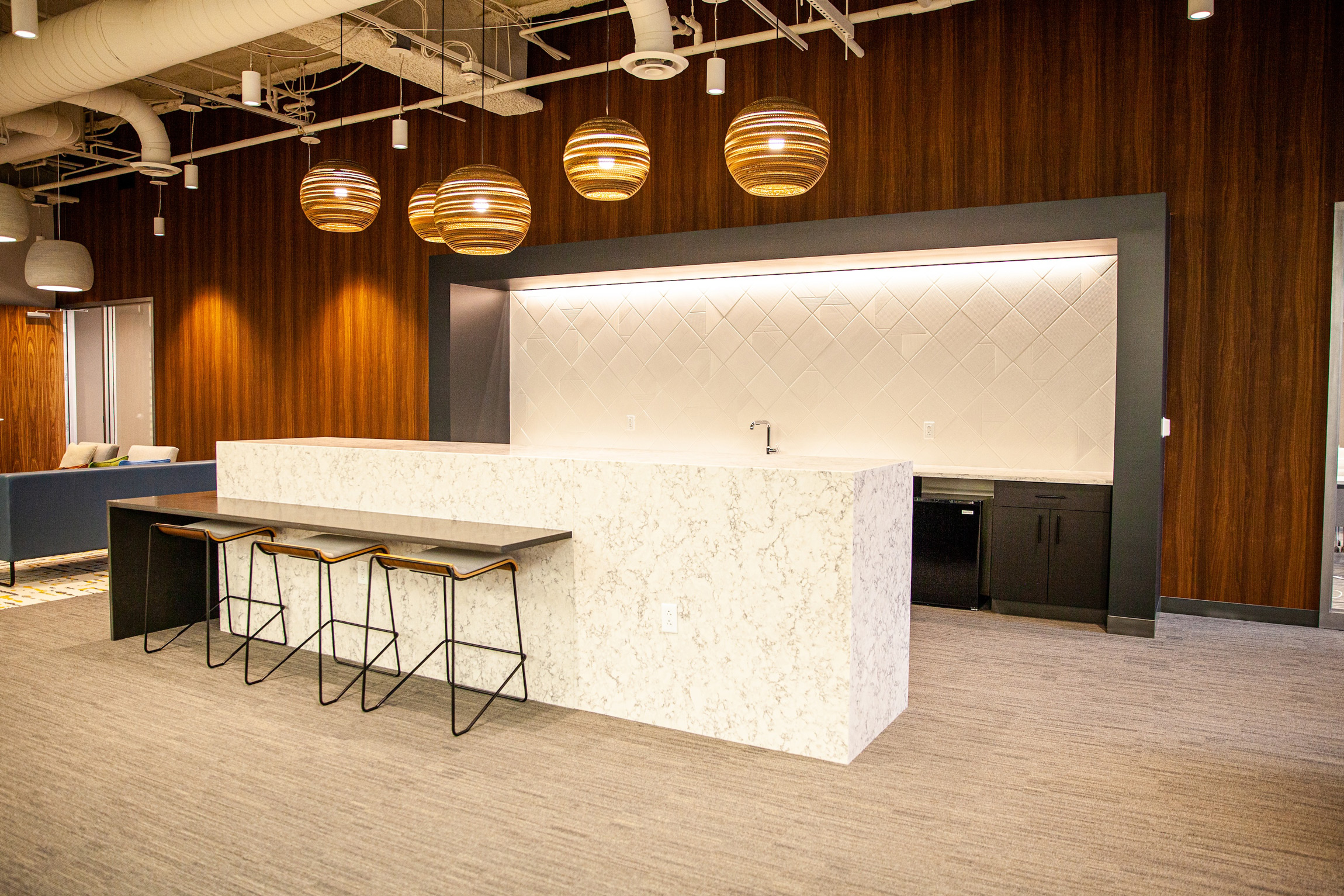 Modern office kitchenette with white marble island, three bar stools, wood-paneled walls, and round hanging pendant lights.