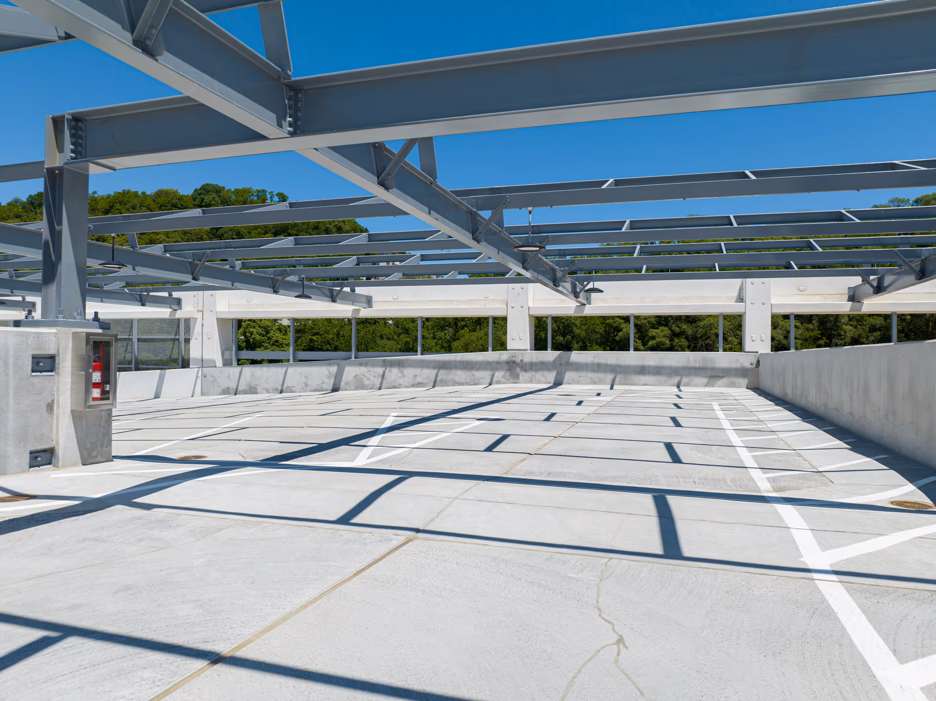 Empty rooftop parking lot with concrete floor and overhead steel beams casting shadows, surrounded by green trees under a clear blue sky.