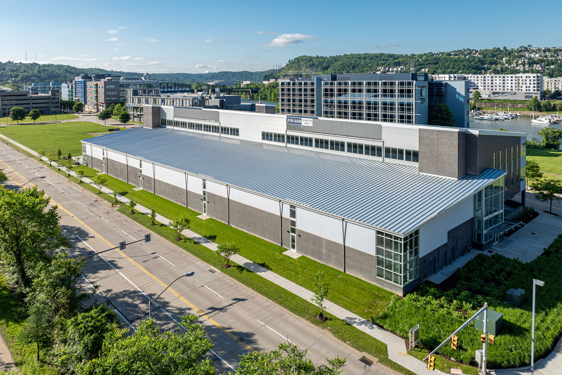 A modern industrial building with a corrugated metal roof and glass windows, surrounded by greenery and adjacent to a road and river.