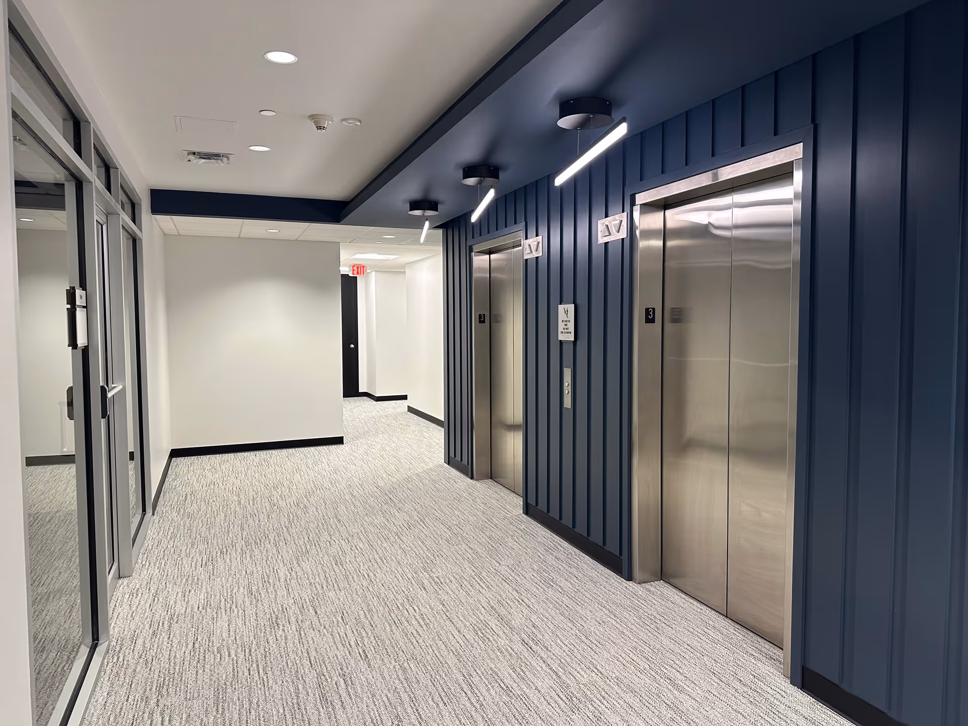 Modern hallway with two closed stainless steel elevators on the right wall and a carpeted floor.