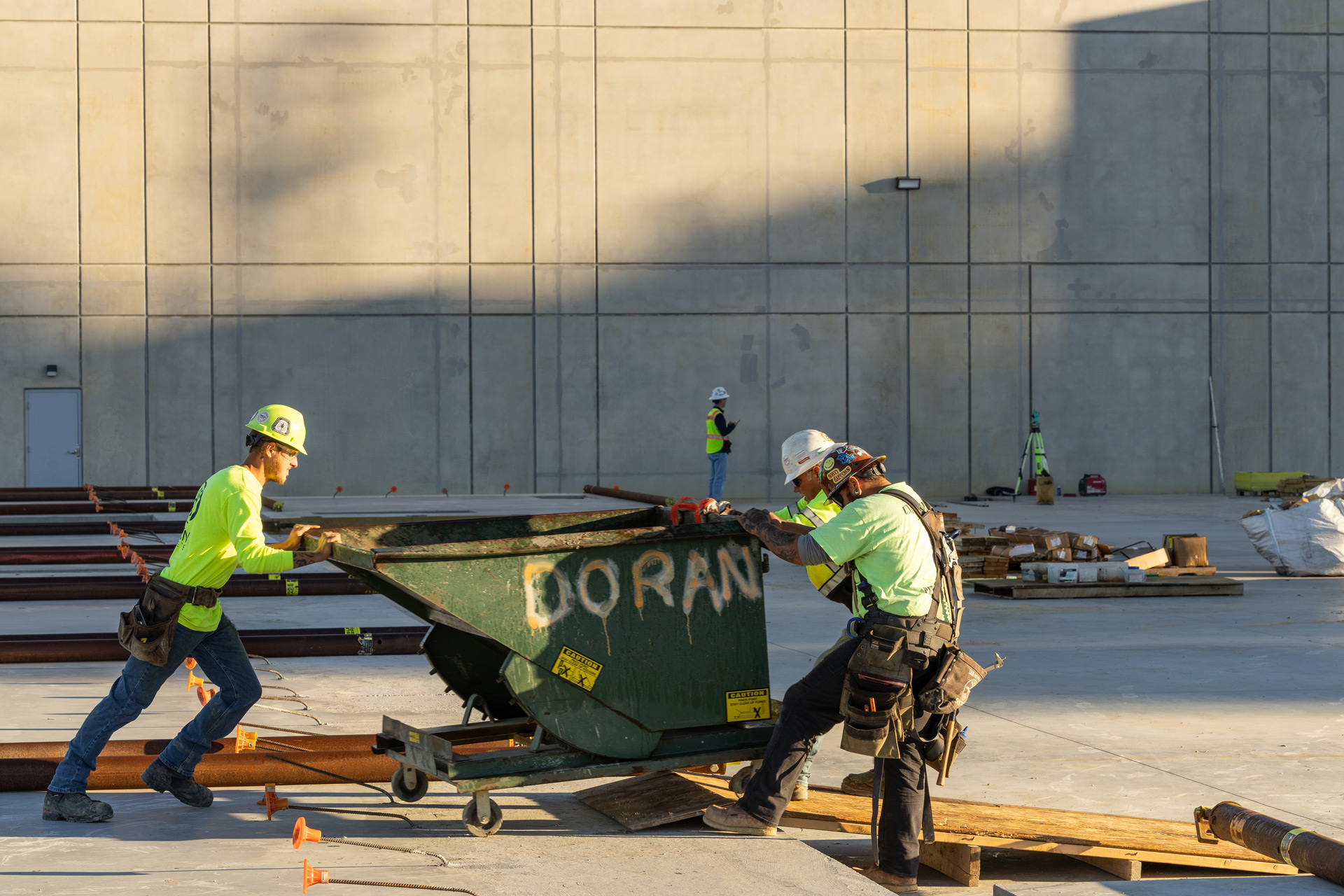 Three construction workers in safety gear pushing a green metal container labeled 'DORAN' on a concrete site with pipes and materials around.