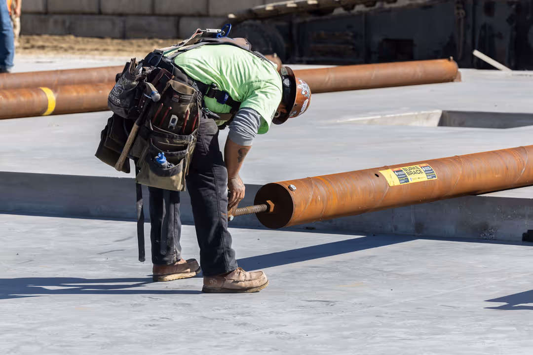 Construction worker in a green shirt and hard hat bending over to inspect a large rusted steel pipe on a concrete surface.