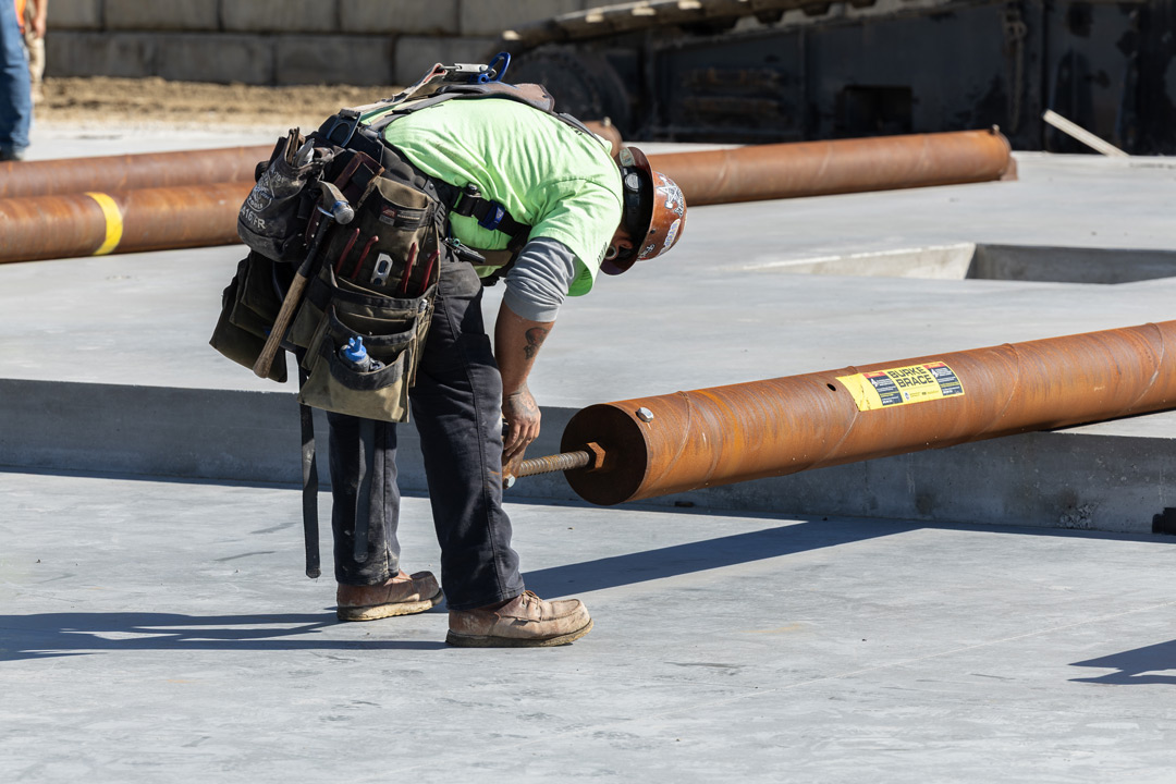 Construction worker in a green shirt and hard hat bending over to inspect a large rusted steel pipe on a concrete surface.