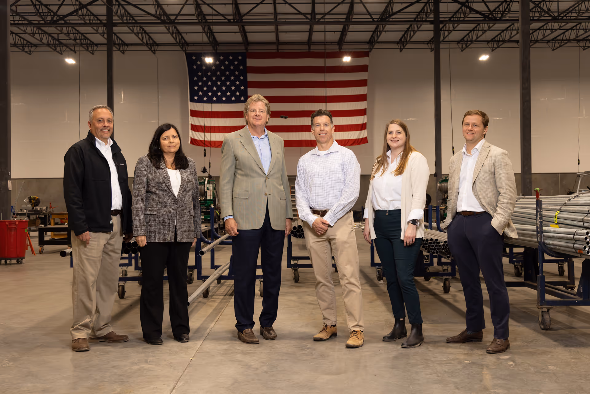 Six professionally dressed people standing inside an industrial warehouse with an American flag hanging on the wall behind them.
