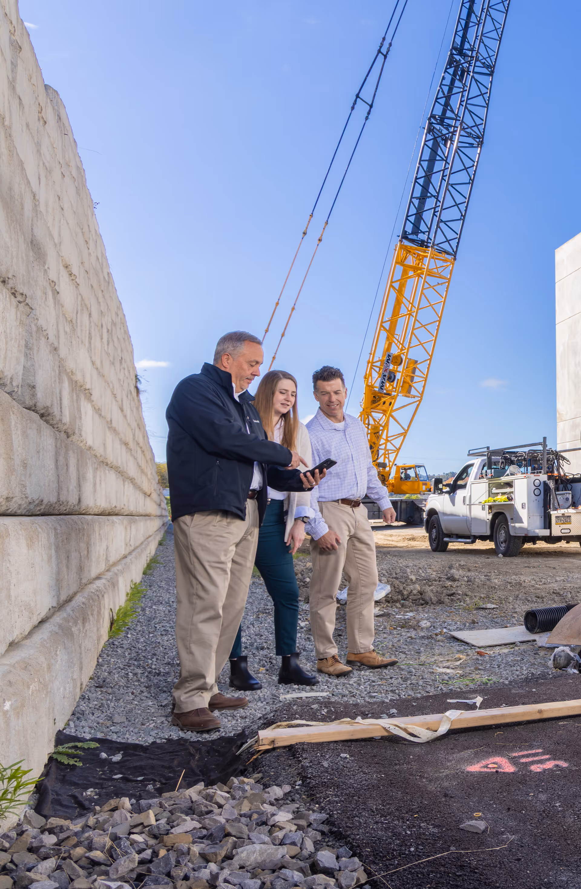 Three people on a construction site looking at a smartphone, with heavy machinery and a truck in the background.