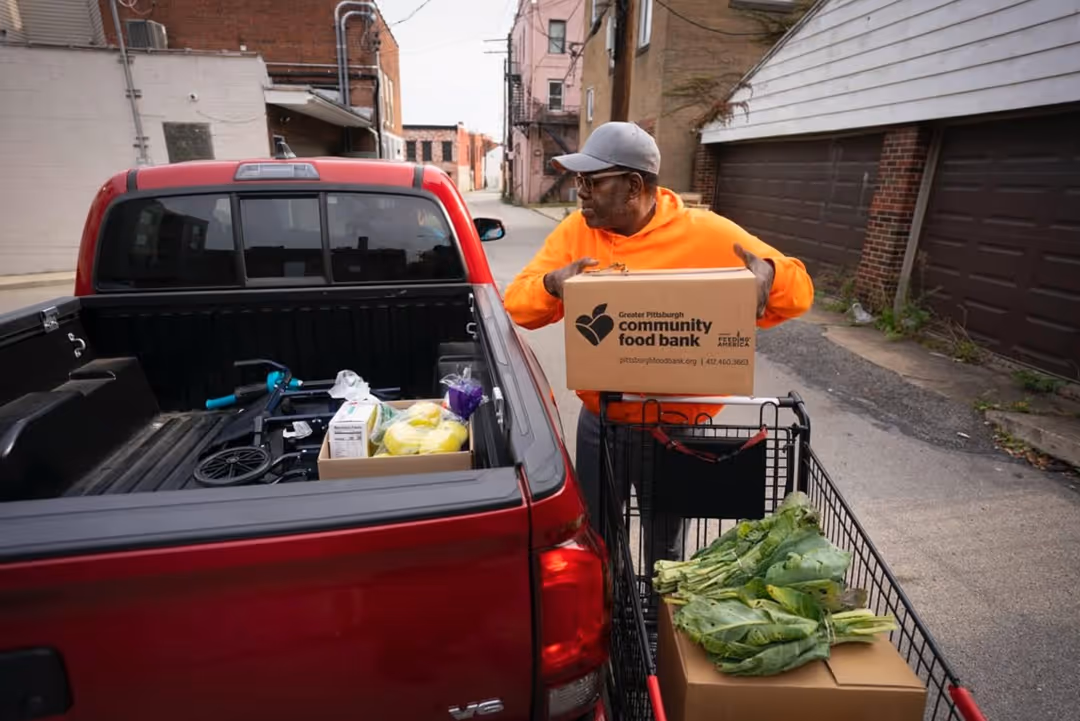 Man in orange hoodie loading a box from Greater Pittsburgh Community Food Bank into a red pickup truck with fresh greens in a shopping cart beside him.