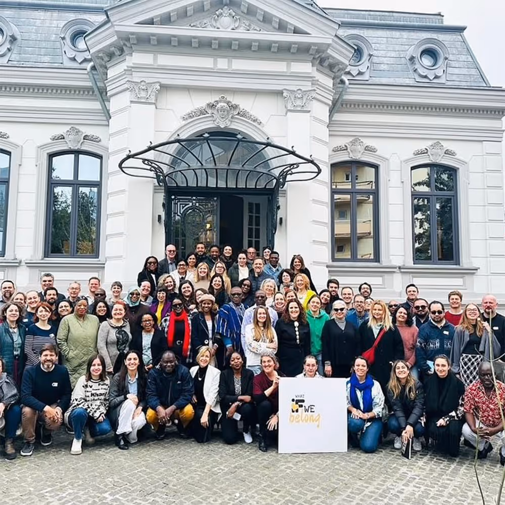 Large diverse group of people posing in front of a white historic building with arched windows and a decorative entrance canopy.