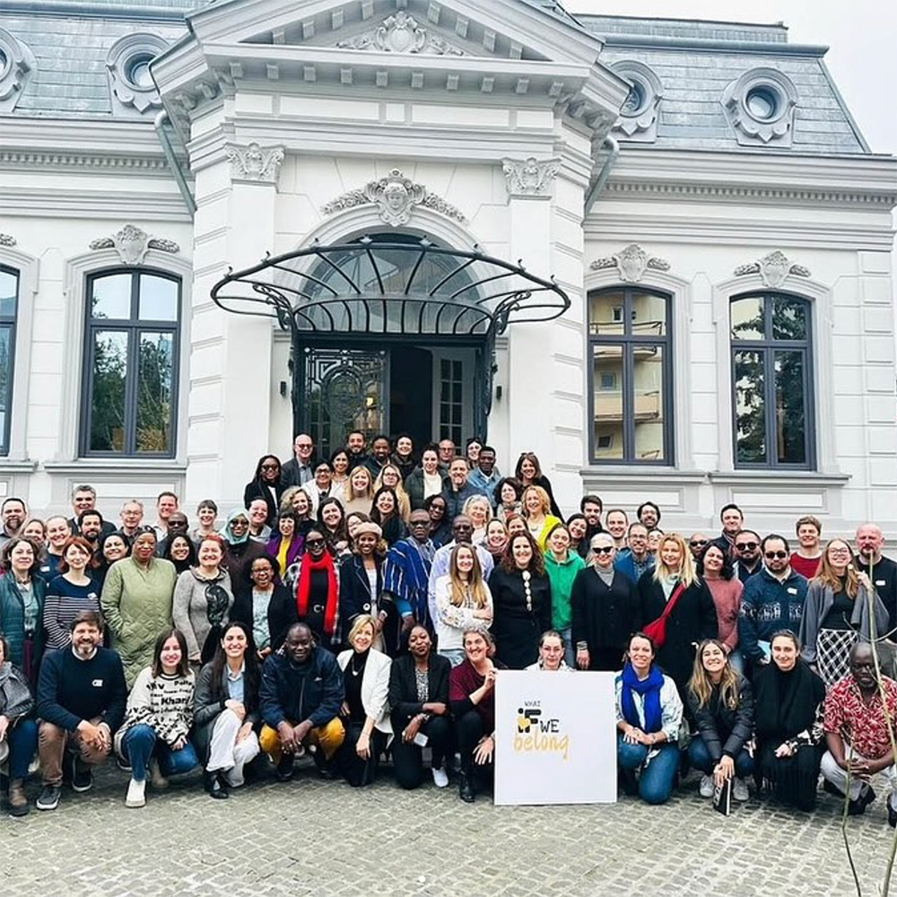 Large diverse group of people posing in front of a white historic building with arched windows and a decorative entrance canopy.
